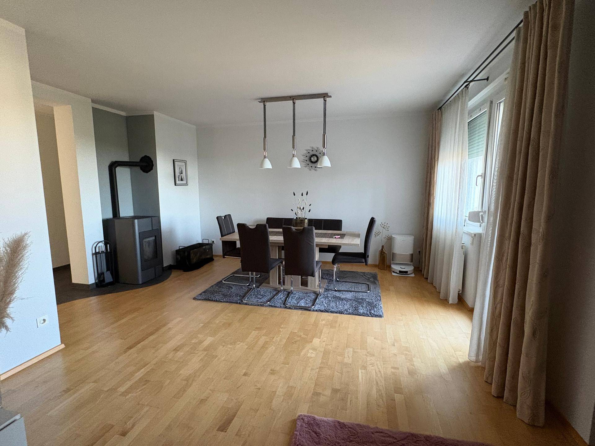 Heller Essbereich mit Holzboden, modernem Kaminofen und großem Fenster mit Vorhängen.