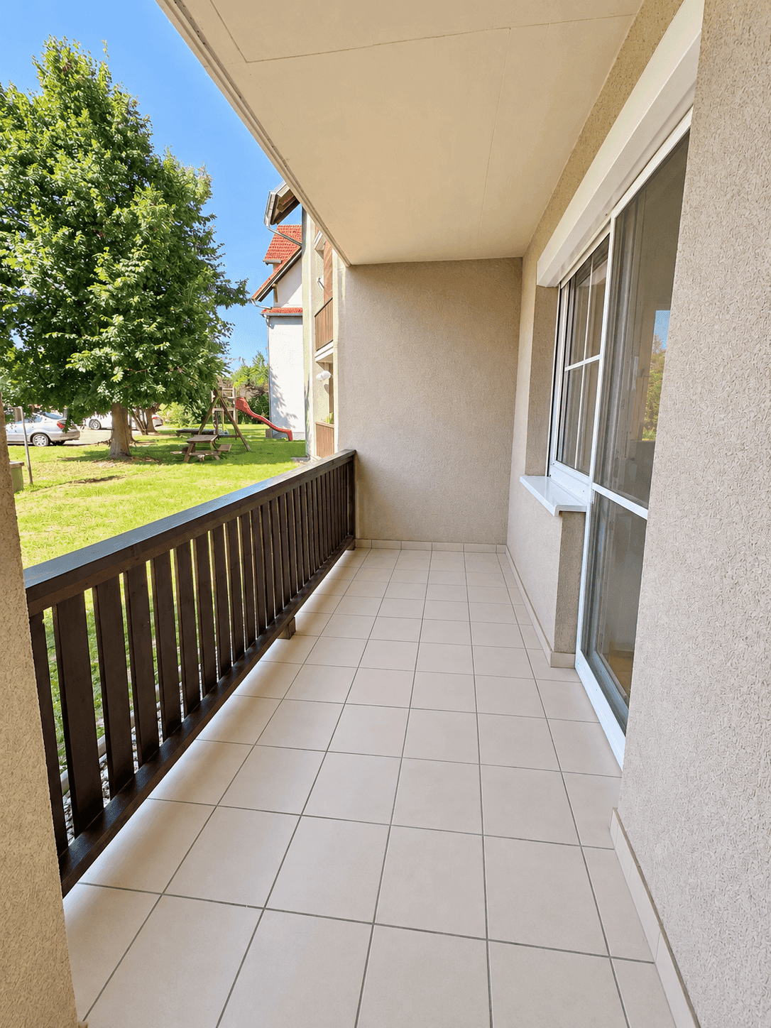 Sonniger Balkon mit Fliesenboden und Blick auf einen Spielplatz und Bäume.
