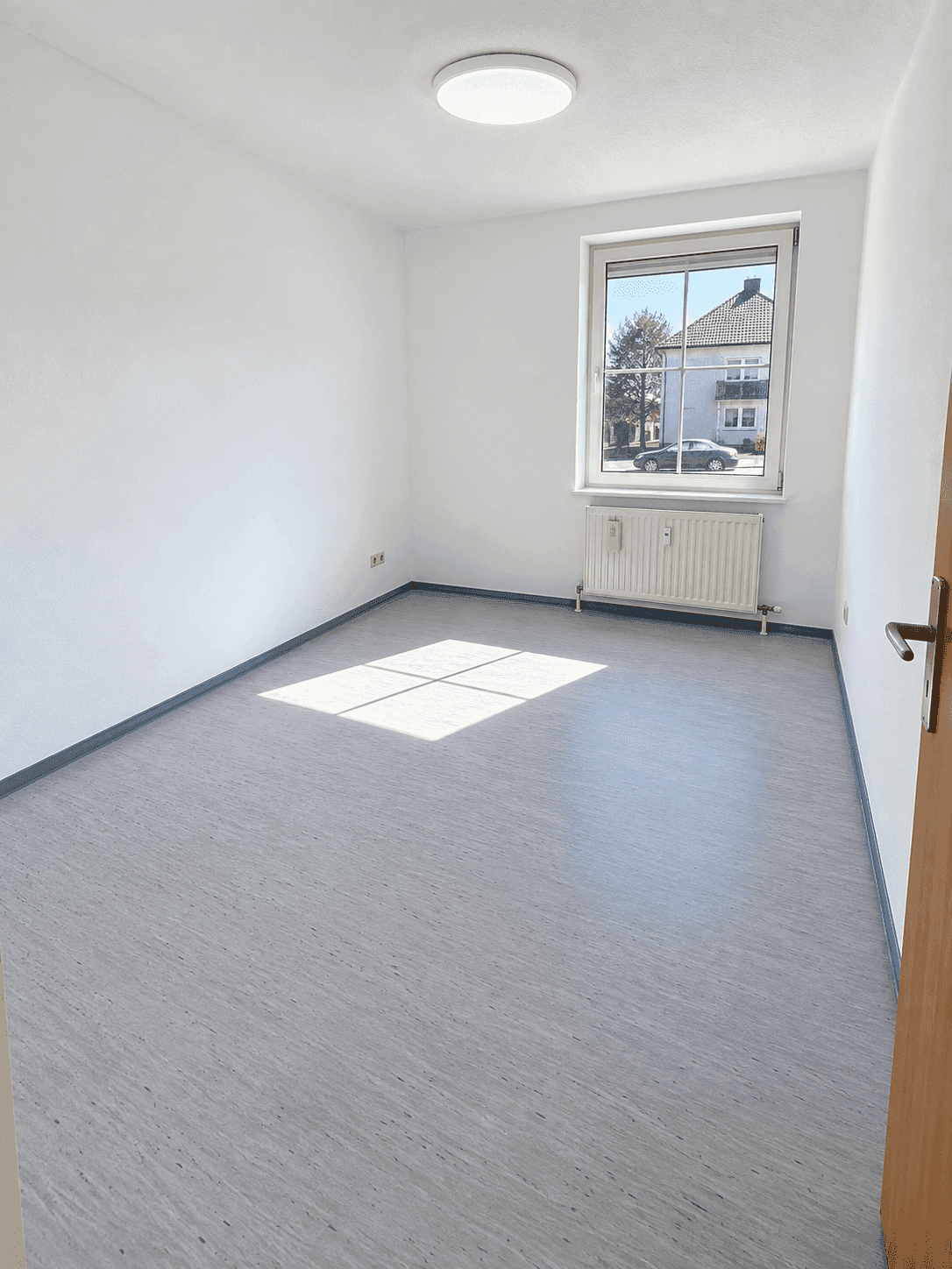 Helles, leeres Zimmer mit grauem Vinylboden und Fensterblick auf die Straße.