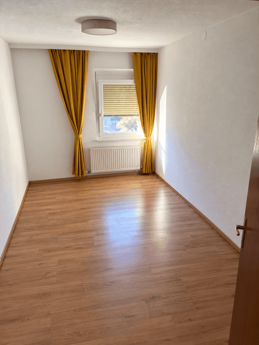 Helles Zimmer mit Laminatboden, Heizkörper und gelben Vorhängen.