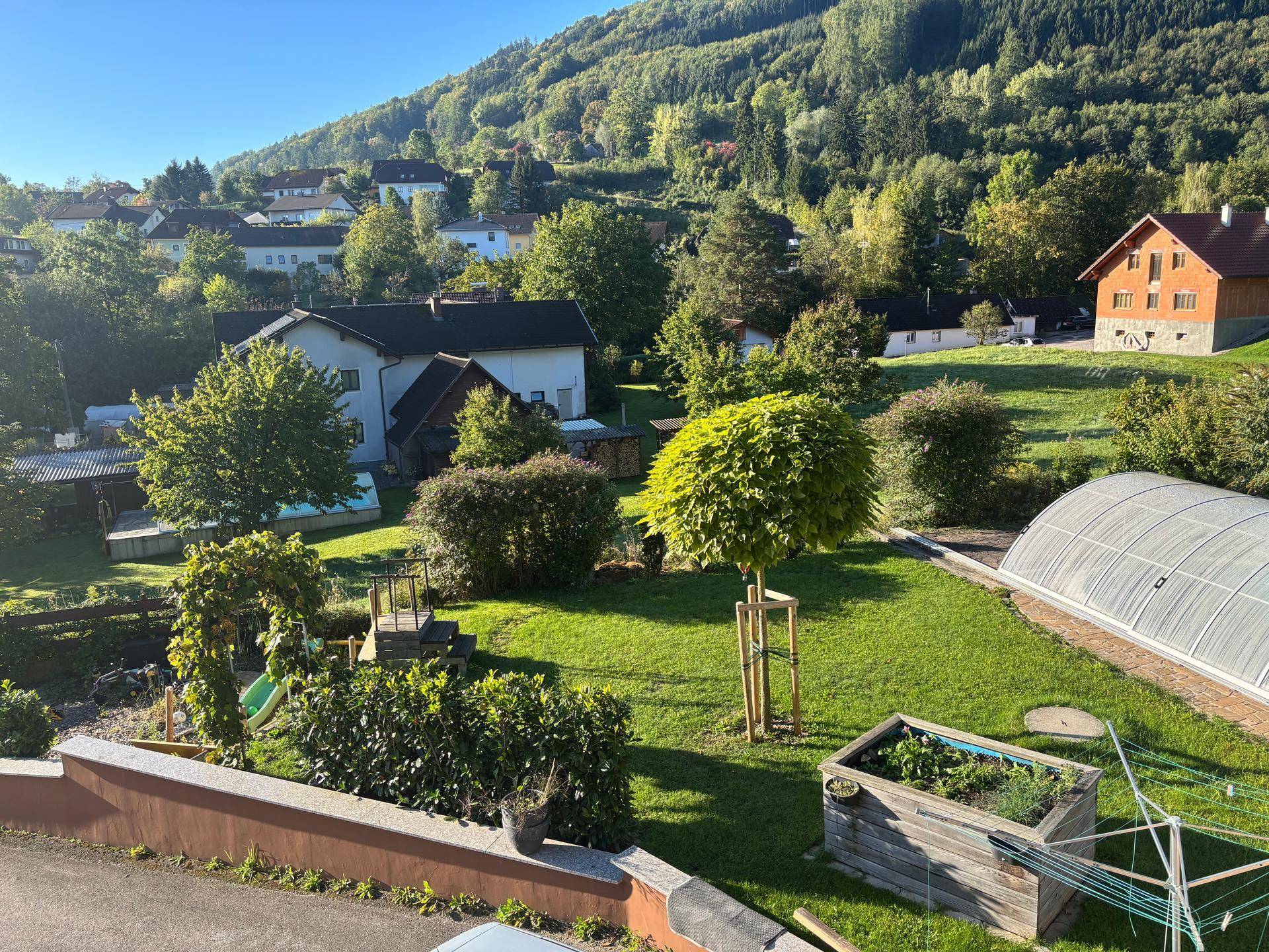 Sonniger Gartenbereich mit Rasenfläche, Hochbeeten und Blick auf die umliegende Hügellandschaft.