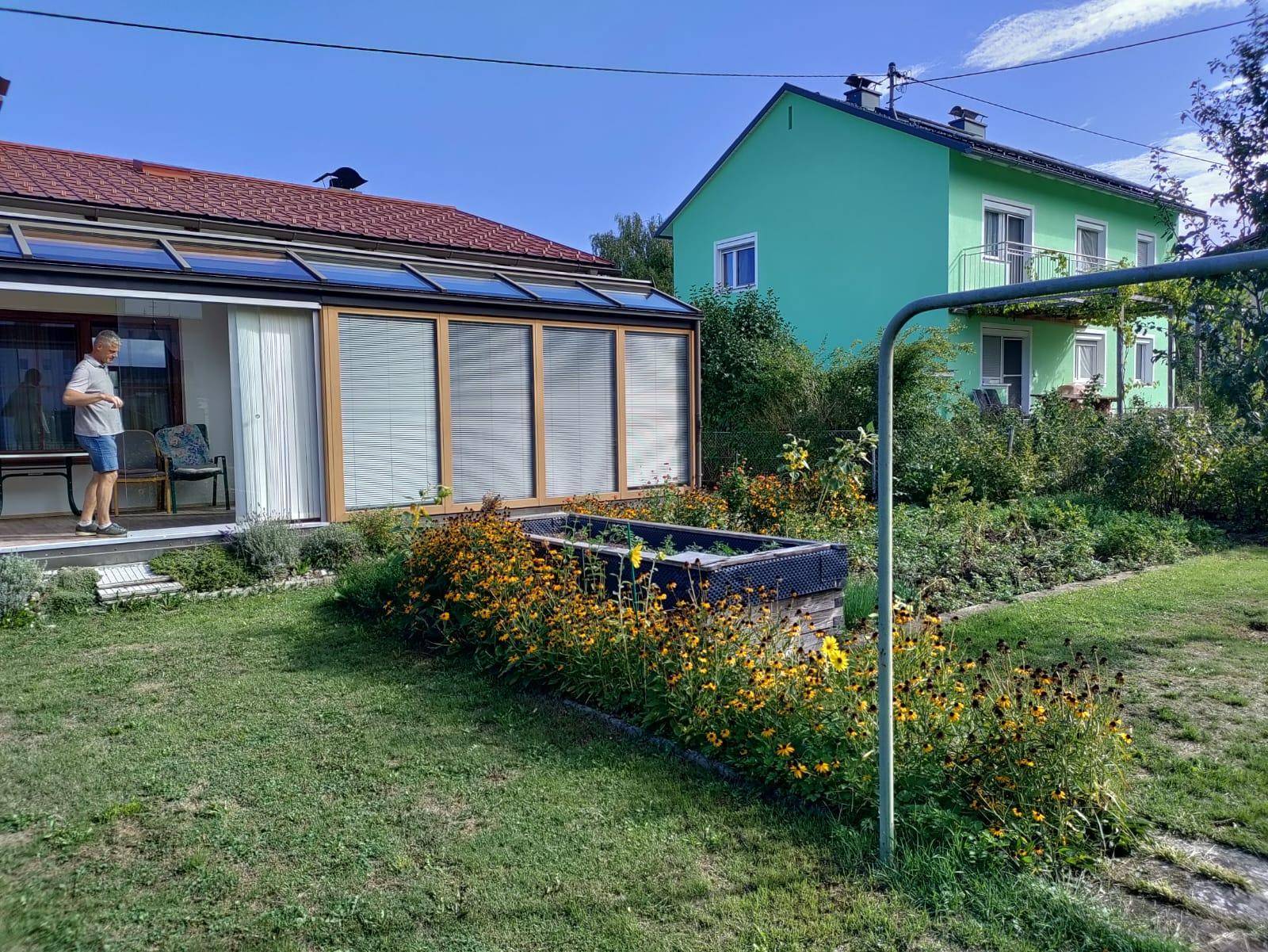 Weitläufiger Garten mit Wintergarten, Blumenbeeten und Blick auf das benachbarte Wohnhaus.