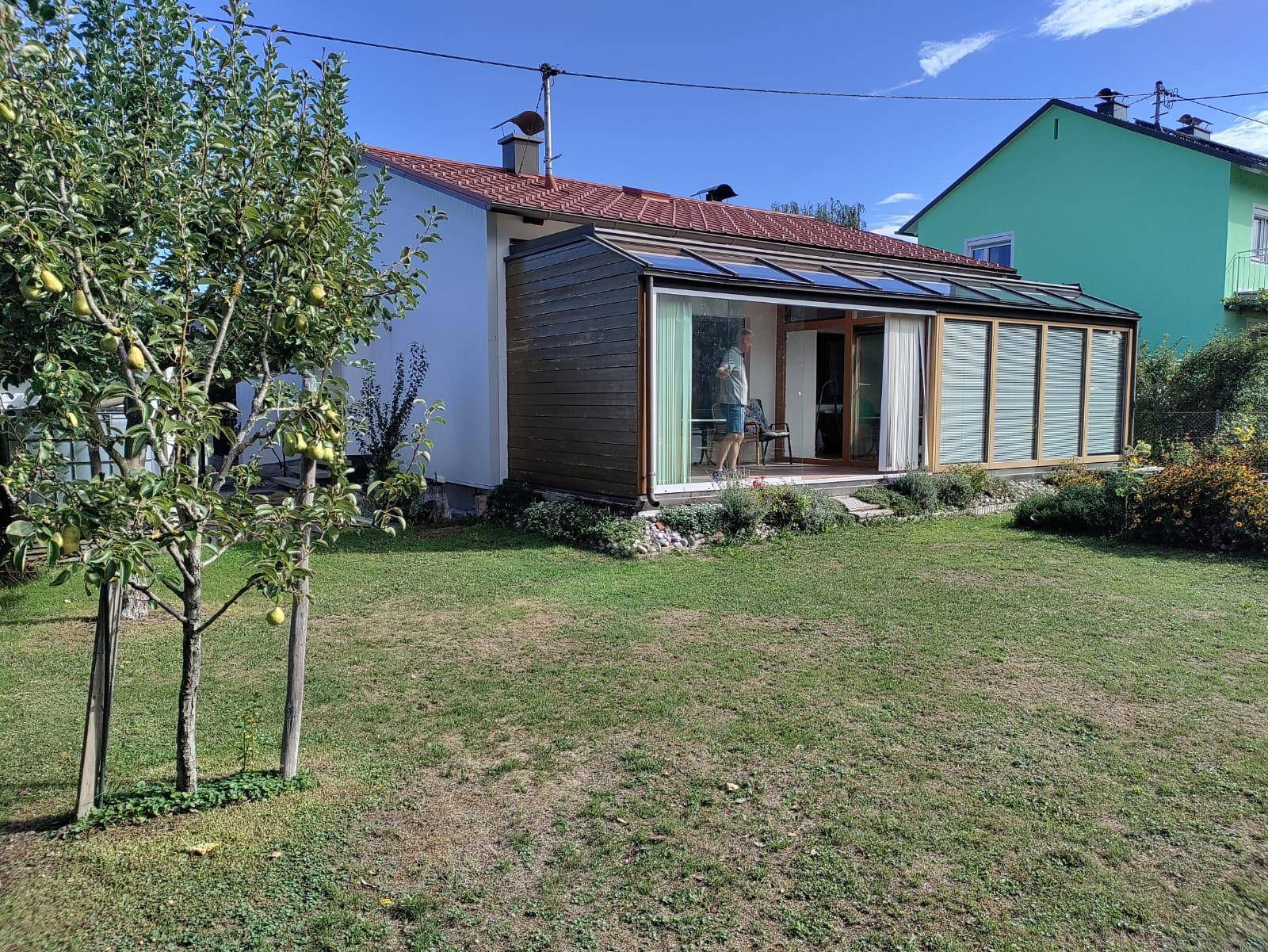 Sonnige Gartenseite des Hauses mit Wintergarten, Rasenfläche und einem jungen Obstbaum.