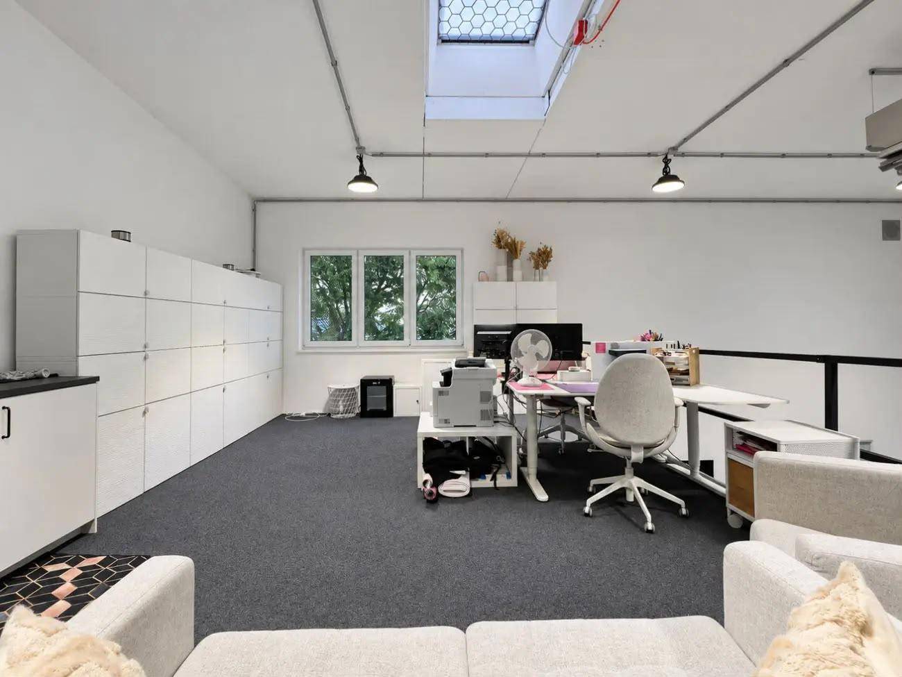 Oberer Bürobereich mit Arbeitsplätzen, Schließfächern und einem Fenster mit Blick ins Grüne.