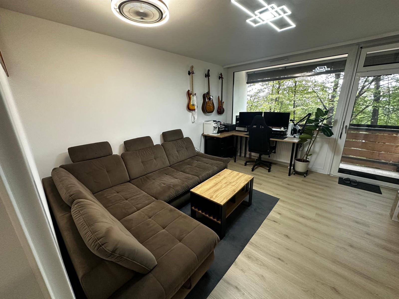 Gemütliches Wohnzimmer mit bequemer Eckcouch und einem Arbeitsplatz am Fenster mit Baumausblick.