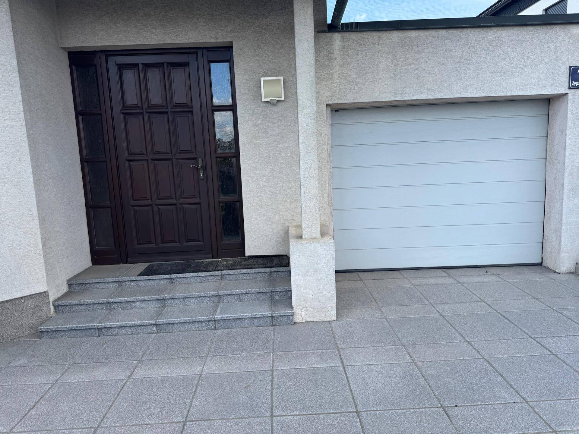 Hauseingang mit dunkler Holztür und integrierter Garage, umgeben von gepflastertem Bereich.