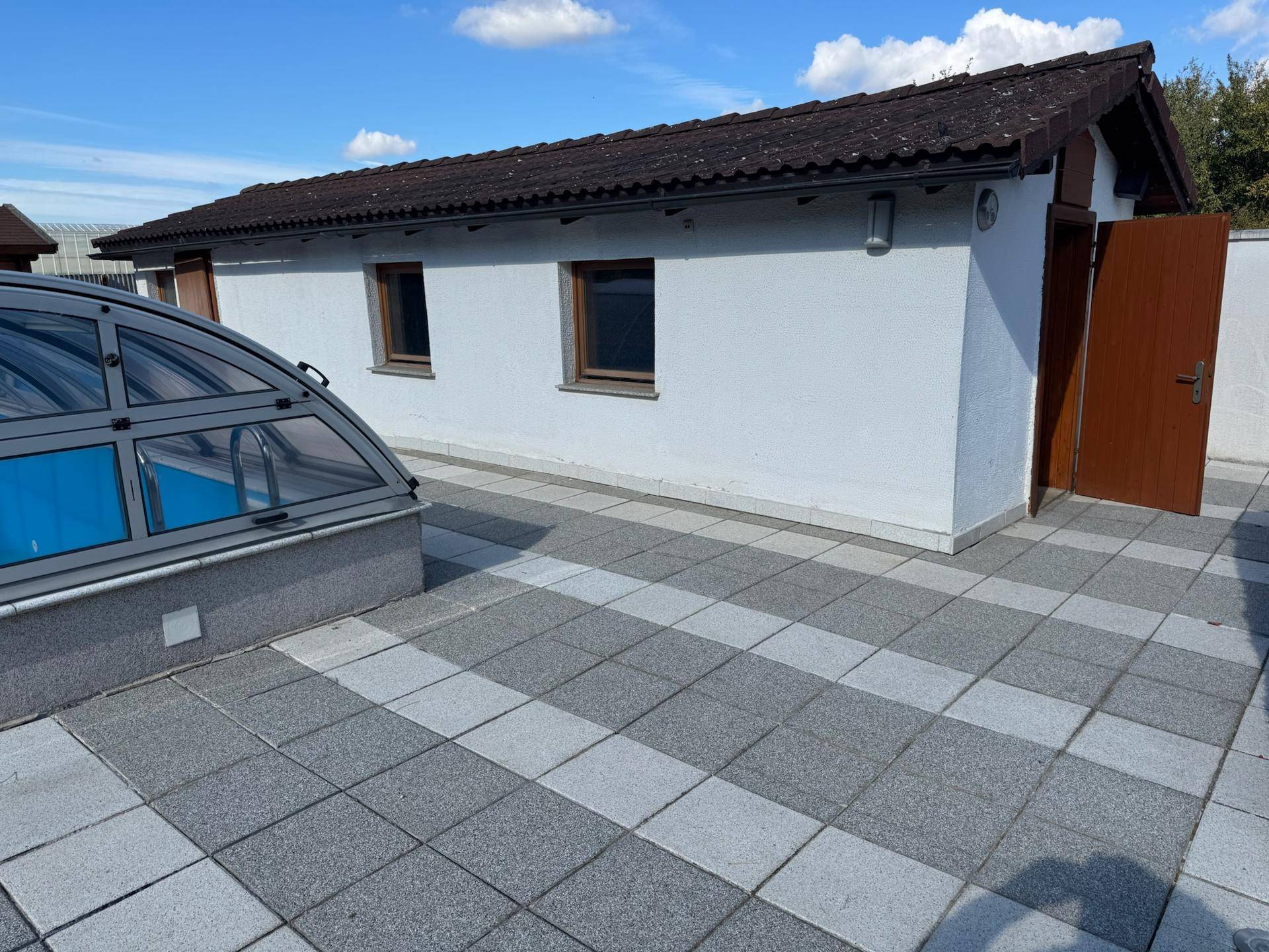 Nebengebäude mit Fenstern und Holztür neben dem Poolbereich auf einer gepflasterten Fläche.