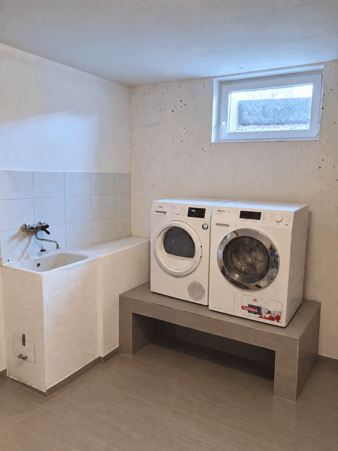 Waschküche im Keller mit Waschmaschine, Trockner, Waschbecken und kleinem Fenster.