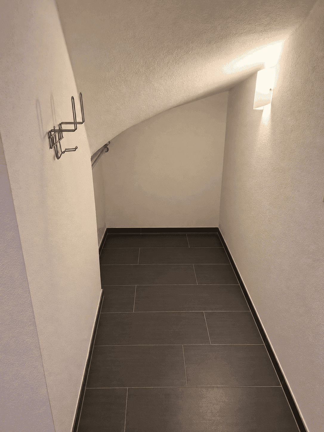 Kleiner Abstellraum unter der Treppe mit dunklen Fliesen und schräger Decke.