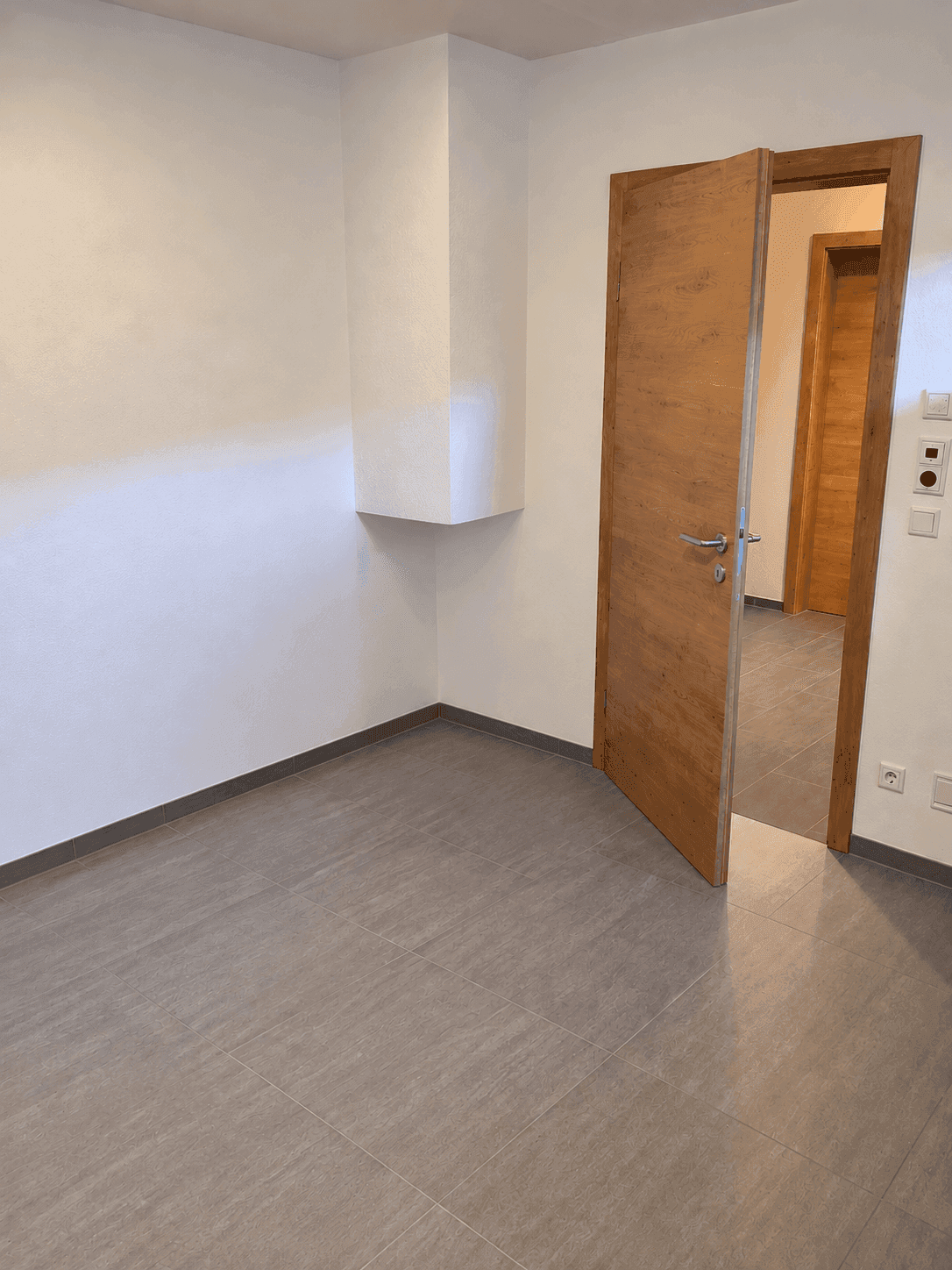 Leerer Raum mit grauen Fliesen, weiß gestrichenen Wänden und Holztür.