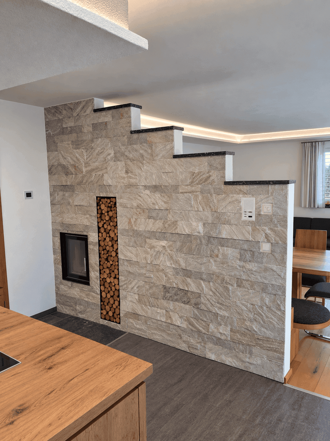 Offener Wohnbereich mit Kamin, Holzstapel und Steinwand, angrenzend an den Essbereich.