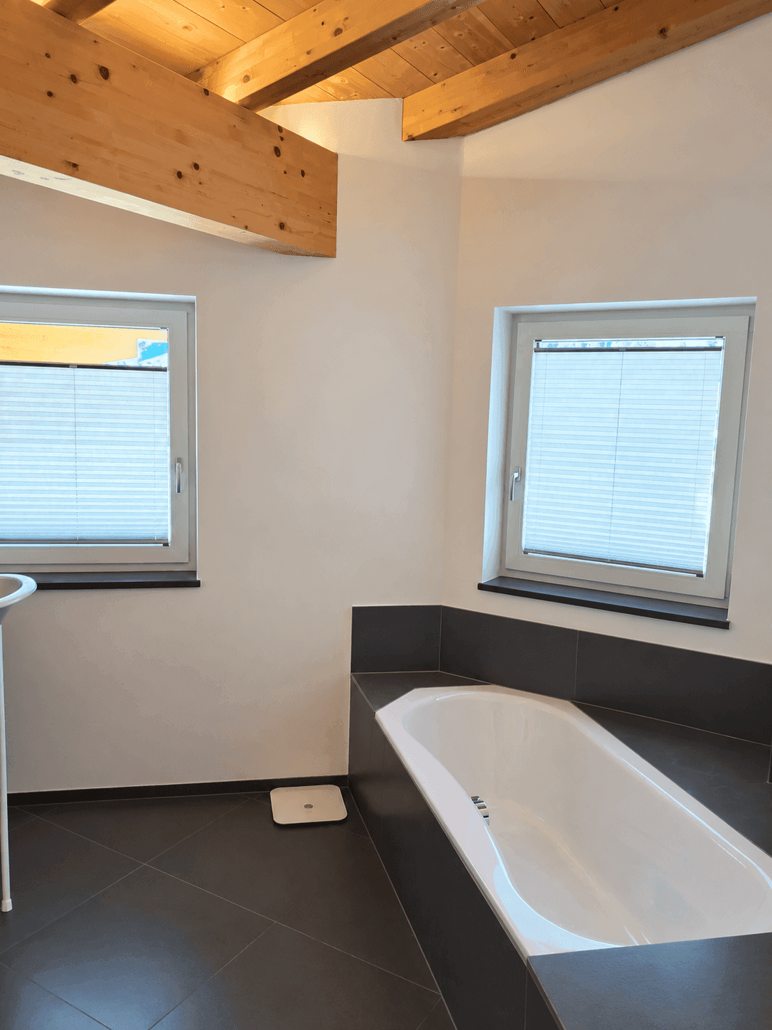 Modernes Badezimmer mit Badewanne, zwei Fenstern und Holzbalkendecke.