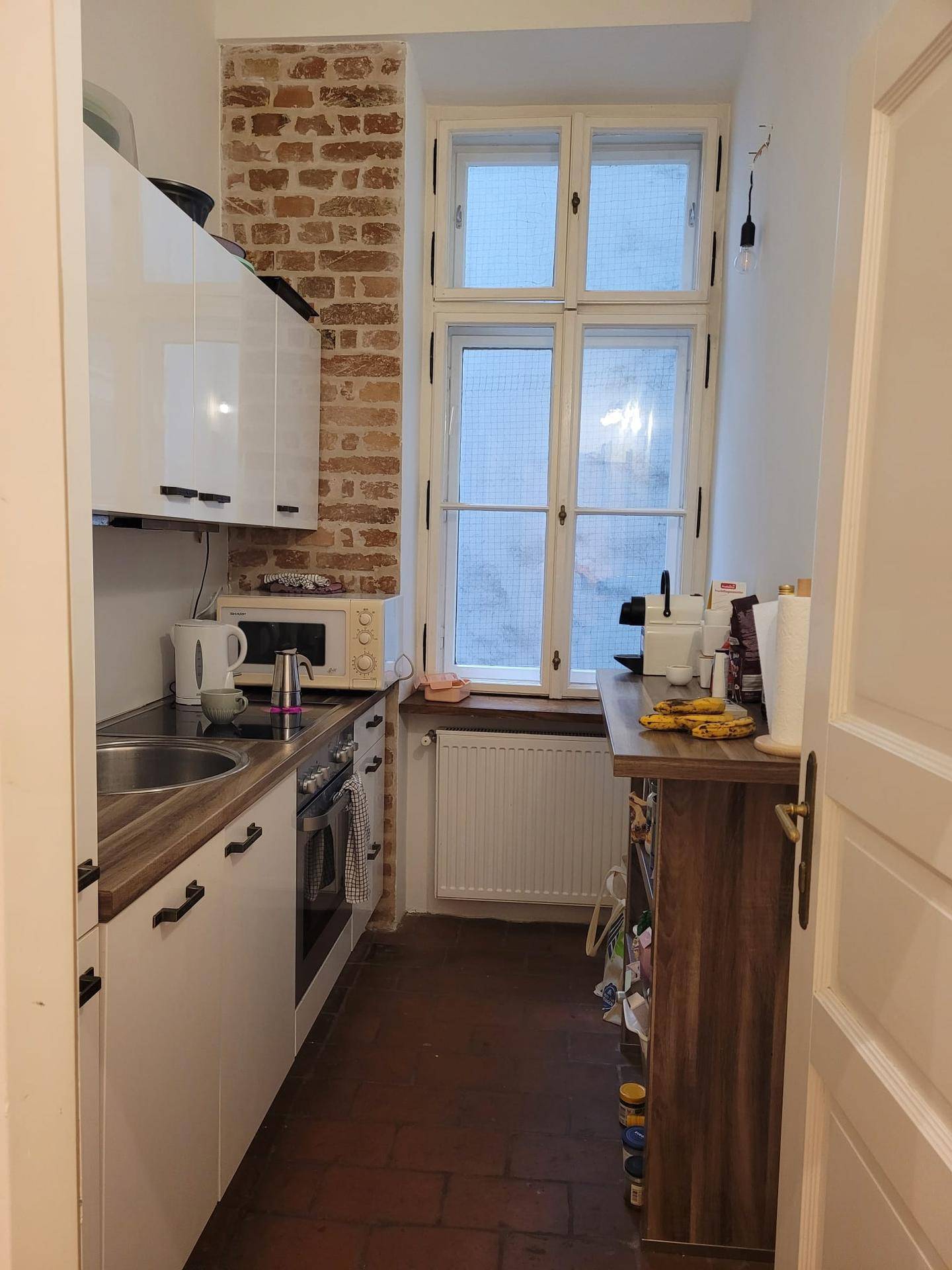 Schmale Küche mit weißen Schränken, Holzarbeitsplatte, freiliegender Ziegelwand und Fenster.