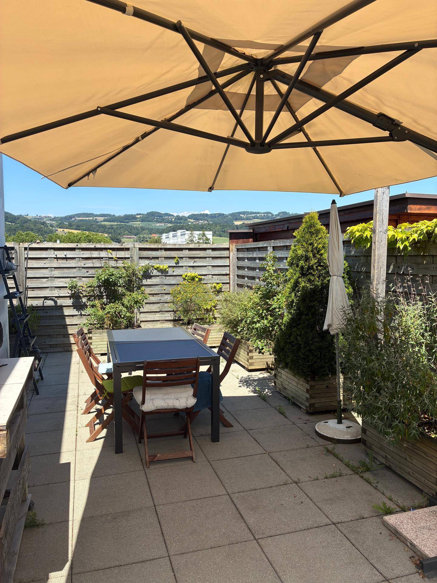 Gemütliche Terrasse mit Esstisch, Stühlen und viel Grün, ideal für Mahlzeiten im Freien unter dem Sonnenschirm.