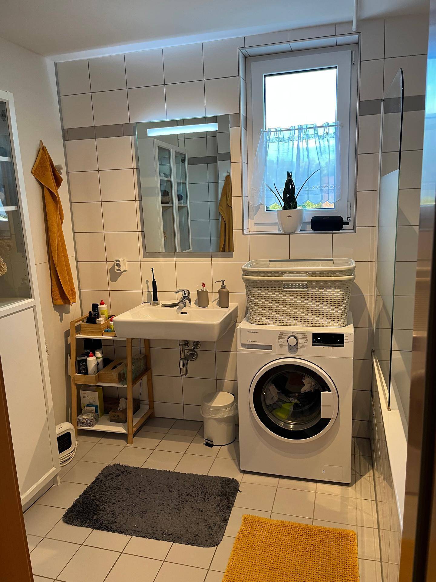 Badezimmer mit Waschbecken, Badewanne, Waschmaschine und Fenster, hell gefliest.