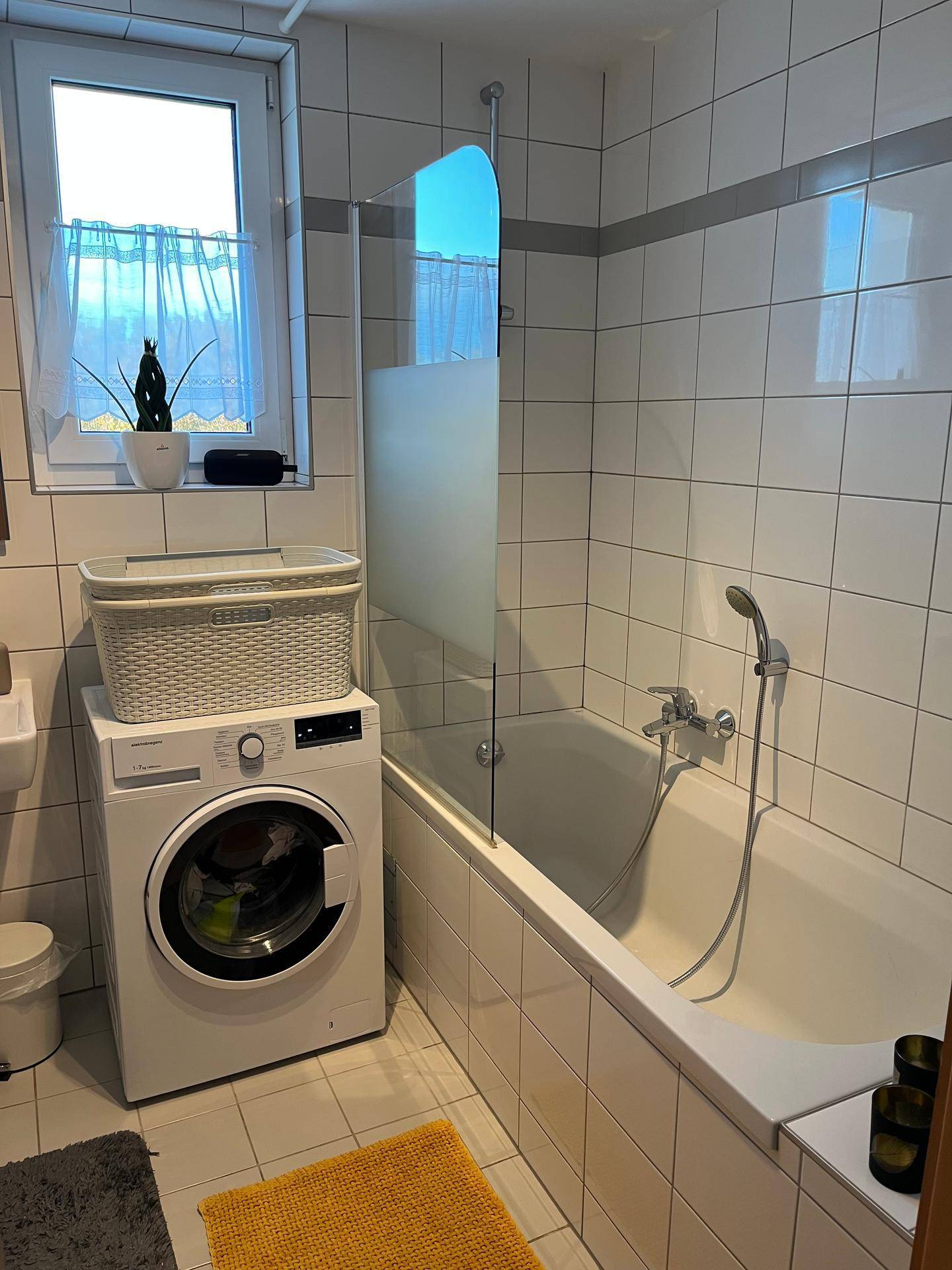Badezimmer mit Badewanne und Duschabtrennung, Waschmaschine und Fenster.