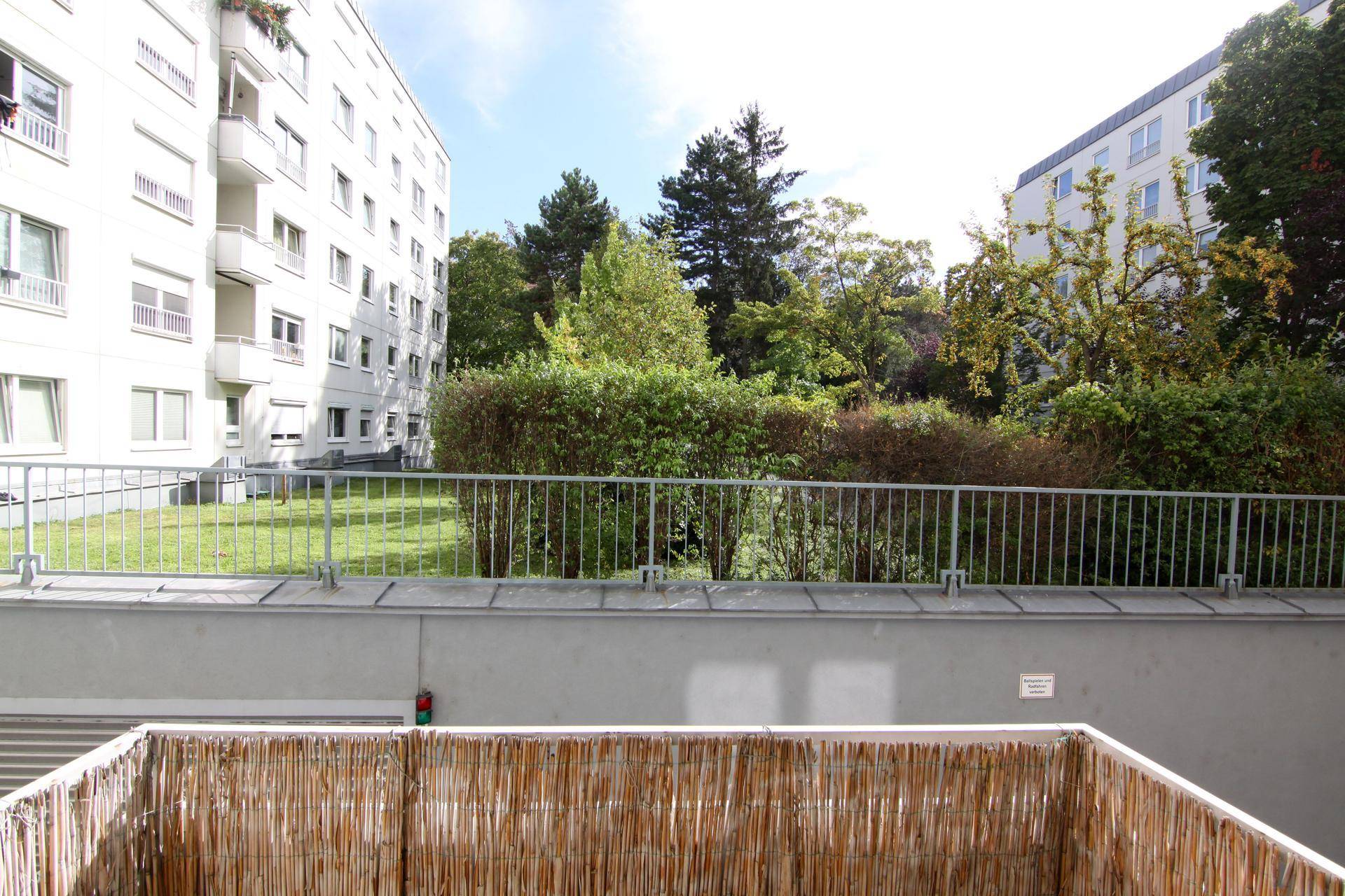 Blick von einem Balkon auf eine grüne Gartenanlage mit Bäumen und umliegenden Gebäuden.