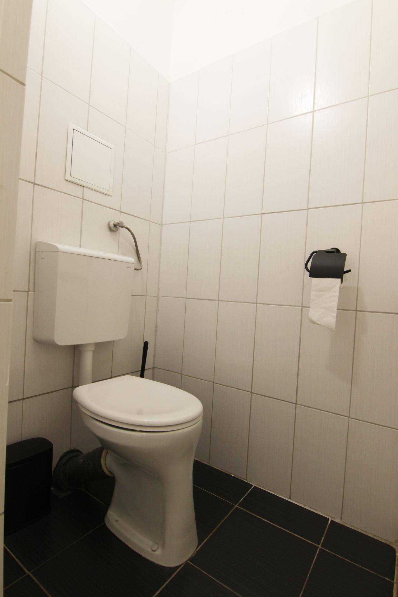 Kleines separates WC mit weißen Wandfliesen und dunklem Boden, funktional und sauber.