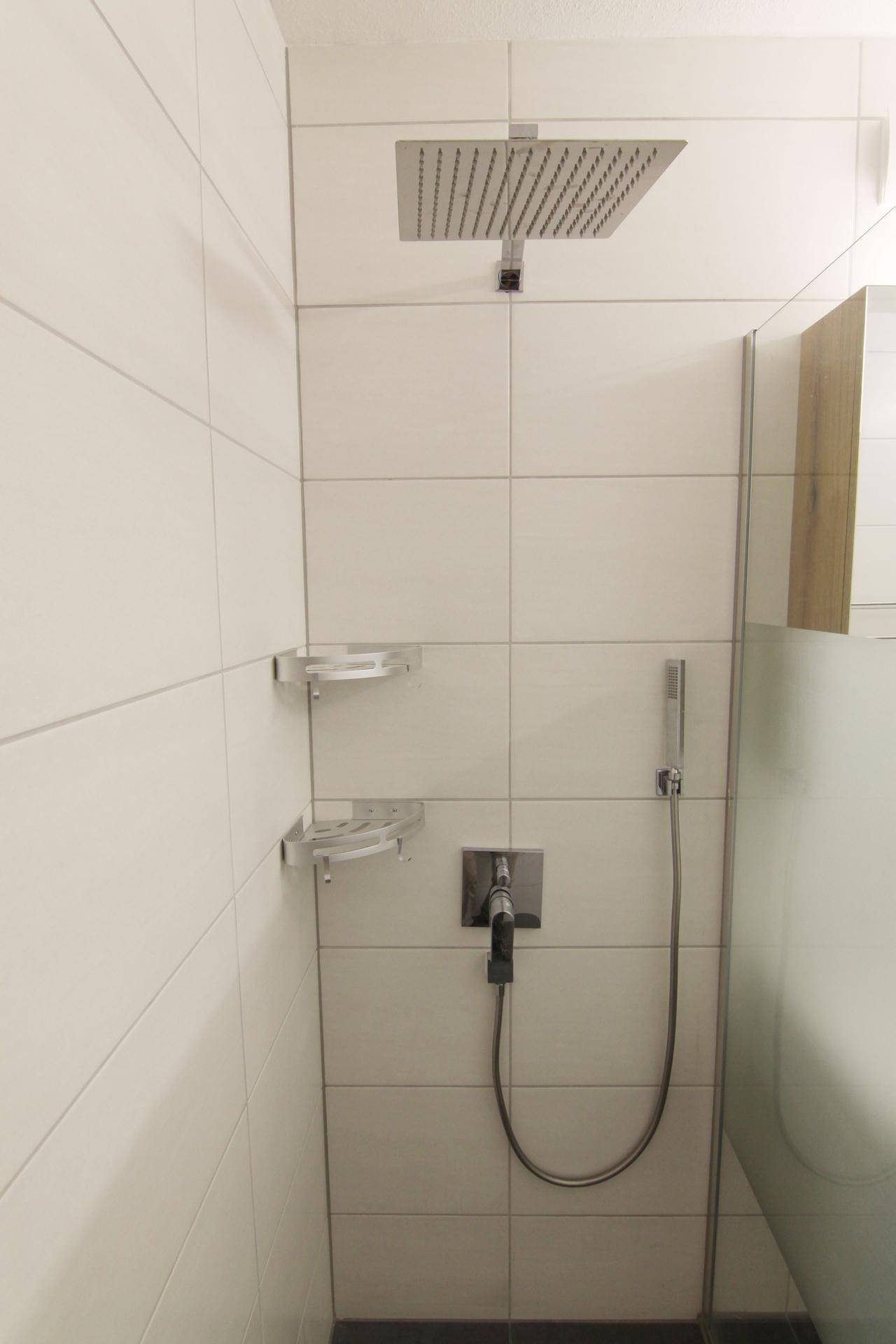 Detailaufnahme der modernen begehbaren Dusche mit Regenduschkopf und weißen Fliesen.