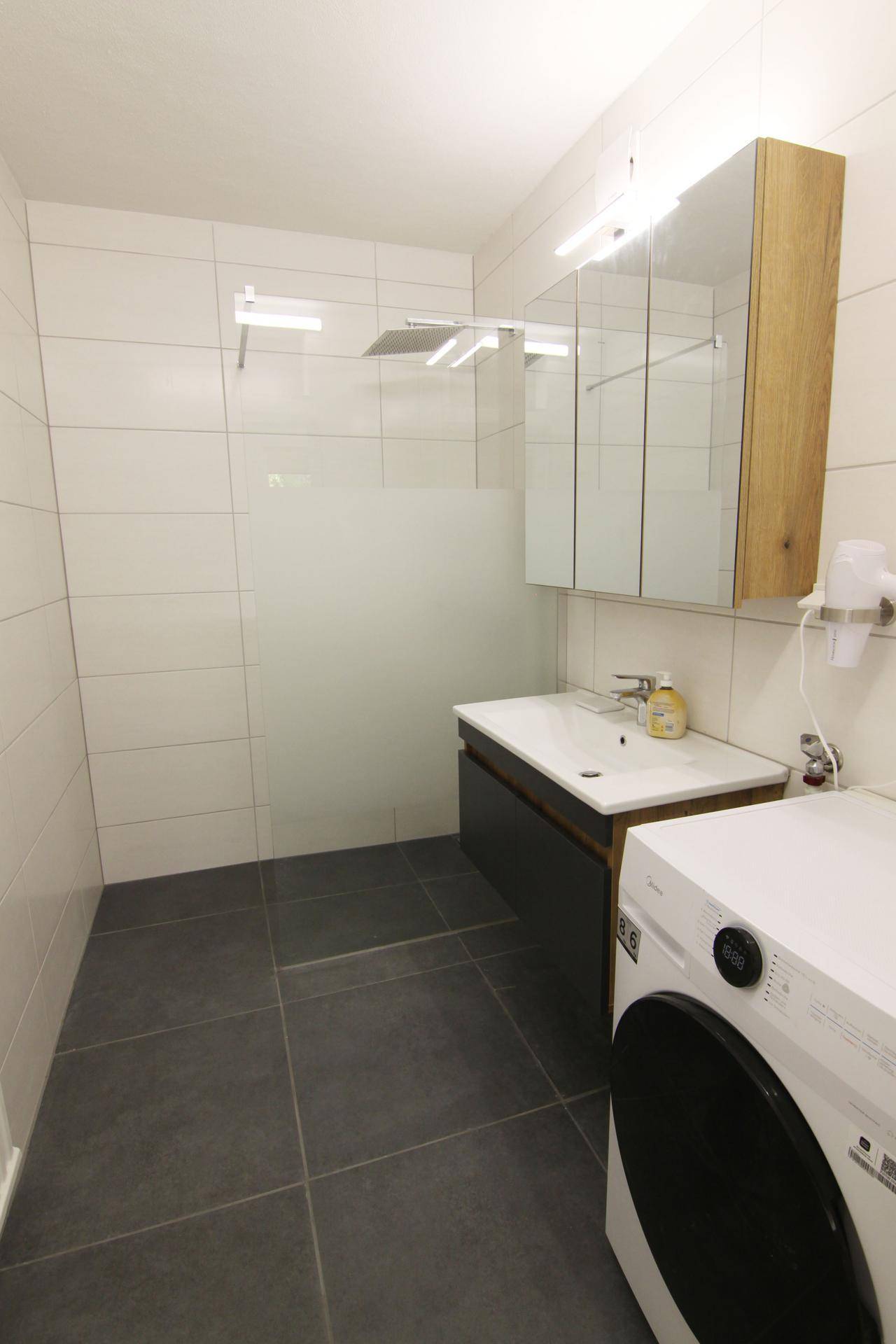 Modernes Badezimmer mit dunklen Fliesen, begehbarer Dusche und Waschmaschine, hell und funktional.