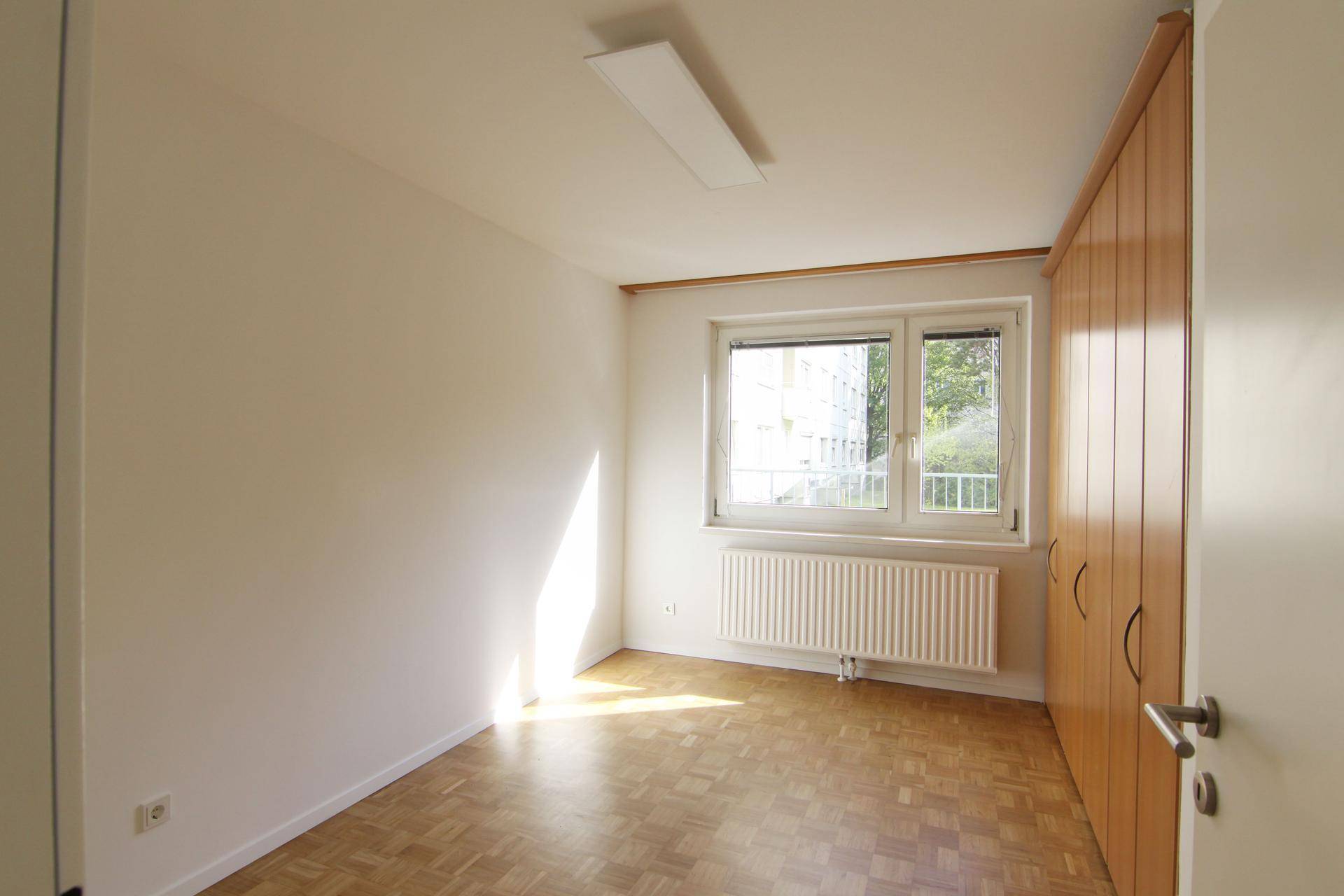 Helles Schlafzimmer mit Parkettboden, Einbauschrank und Fenster mit Blick ins Grüne.