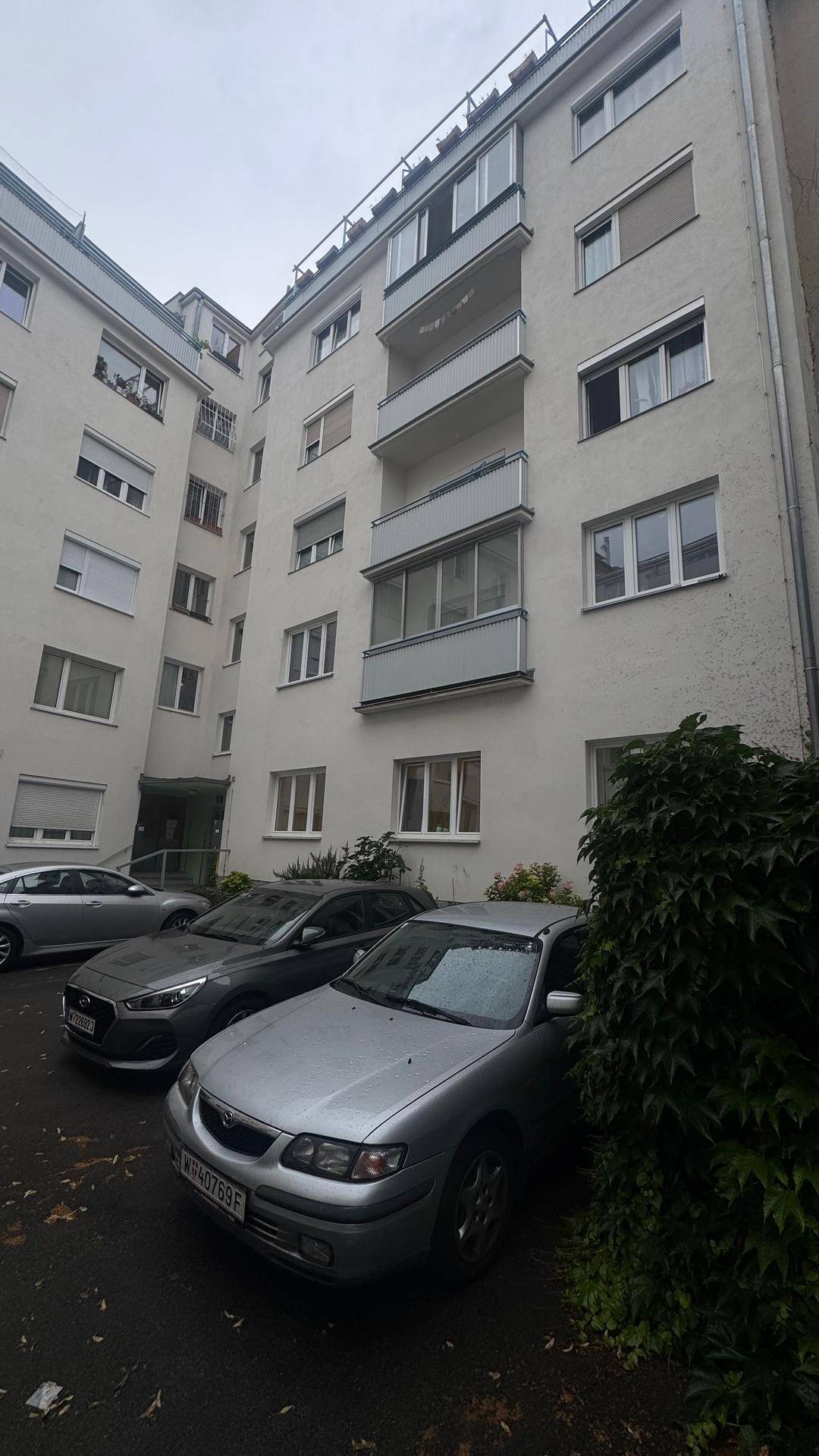 Fassade eines Mehrfamilienhauses mit Balkonen und geparkten Fahrzeugen im Vordergrund.