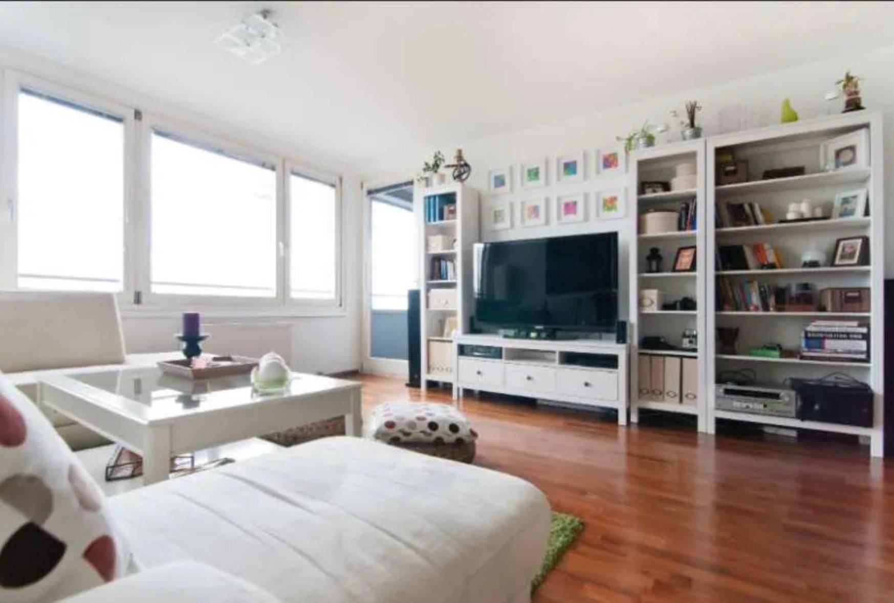 Helles Wohnzimmer mit großen Fenstern, Holzboden und weißer Einrichtung.