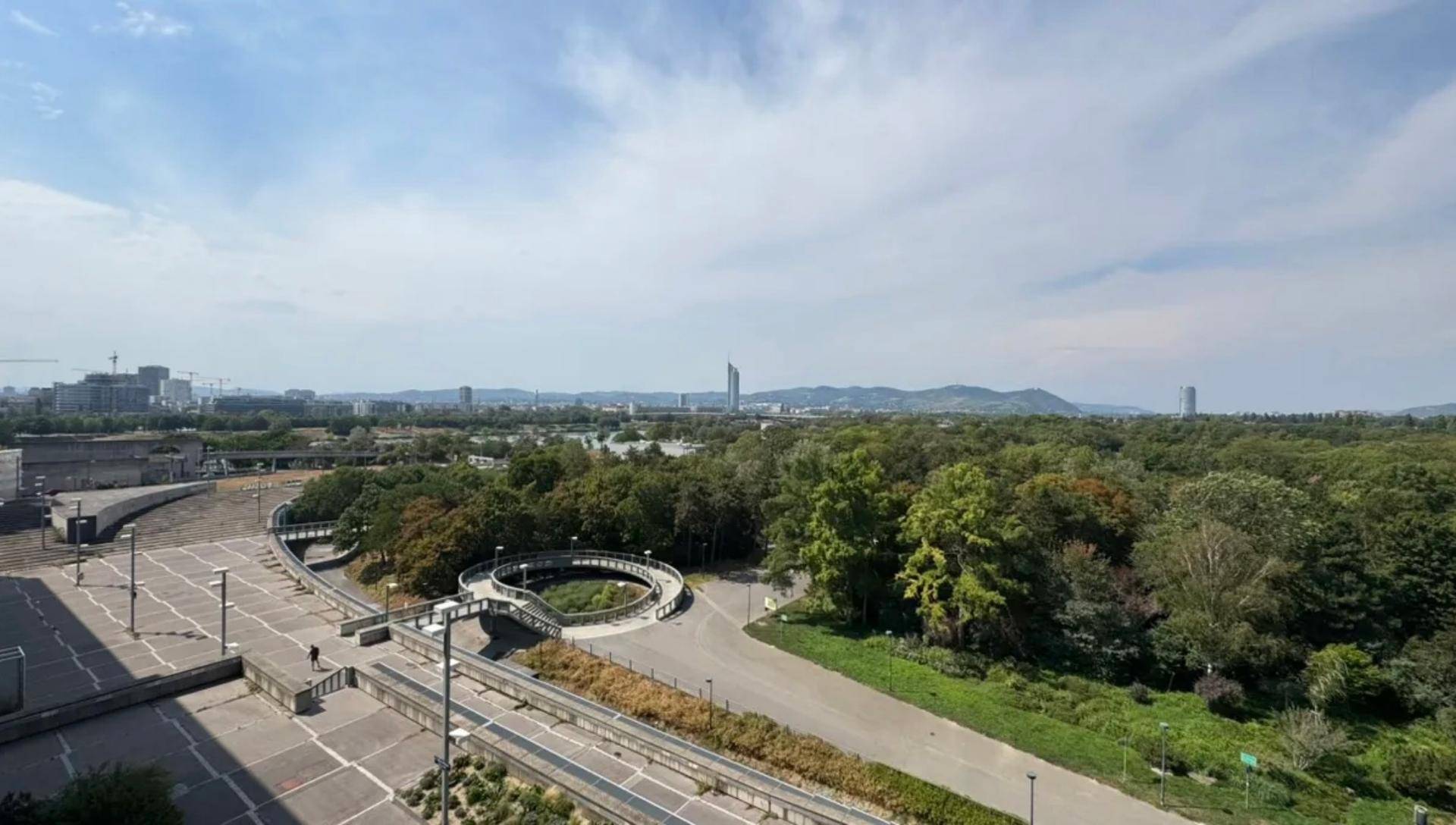 Weitläufiger Blick auf die Stadtlandschaft mit Parkanlagen, Fluss und Bergen im Hintergrund.