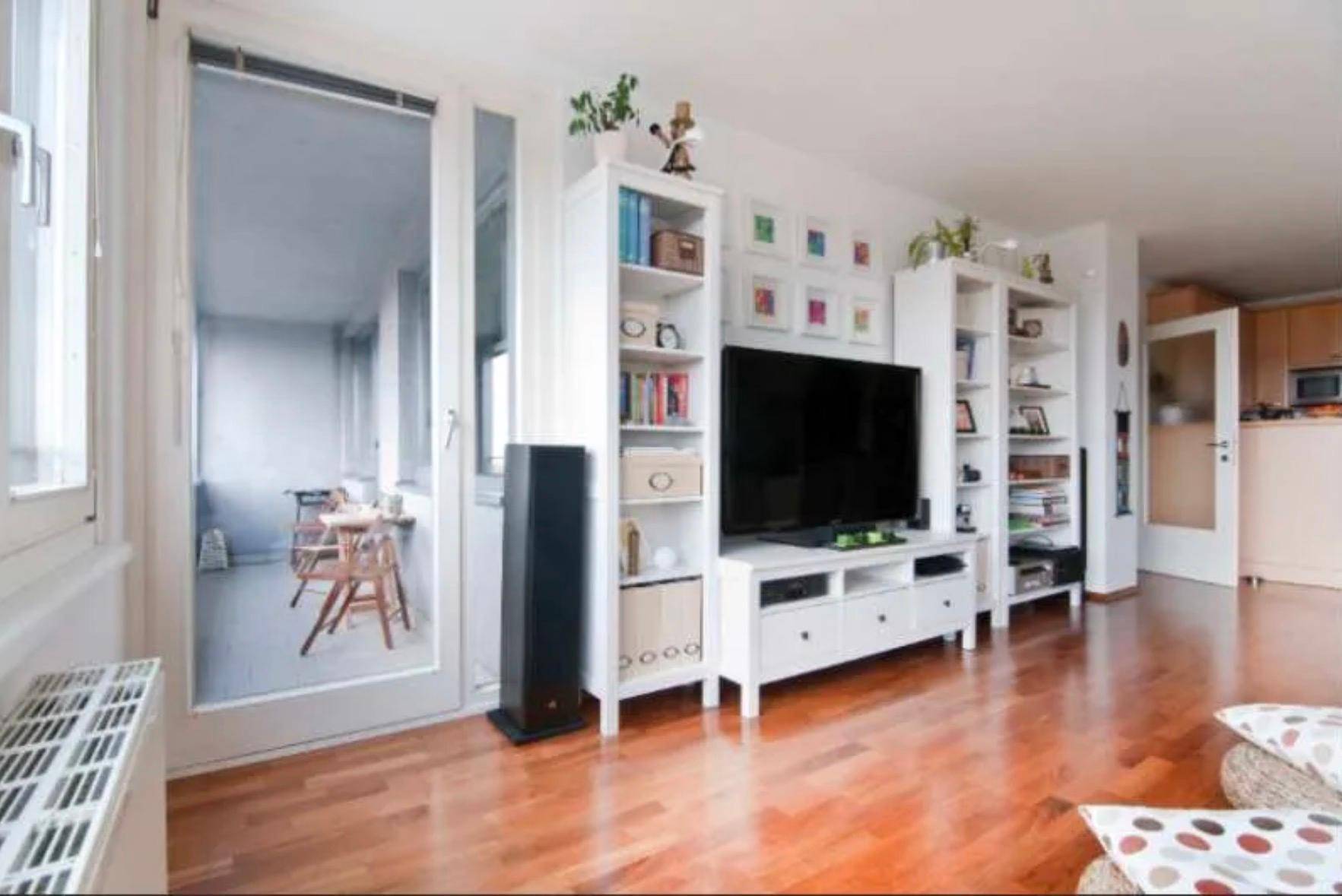 Wohnzimmer mit Holzboden, Bücherregalen und Zugang zu einem verglasten Balkon.