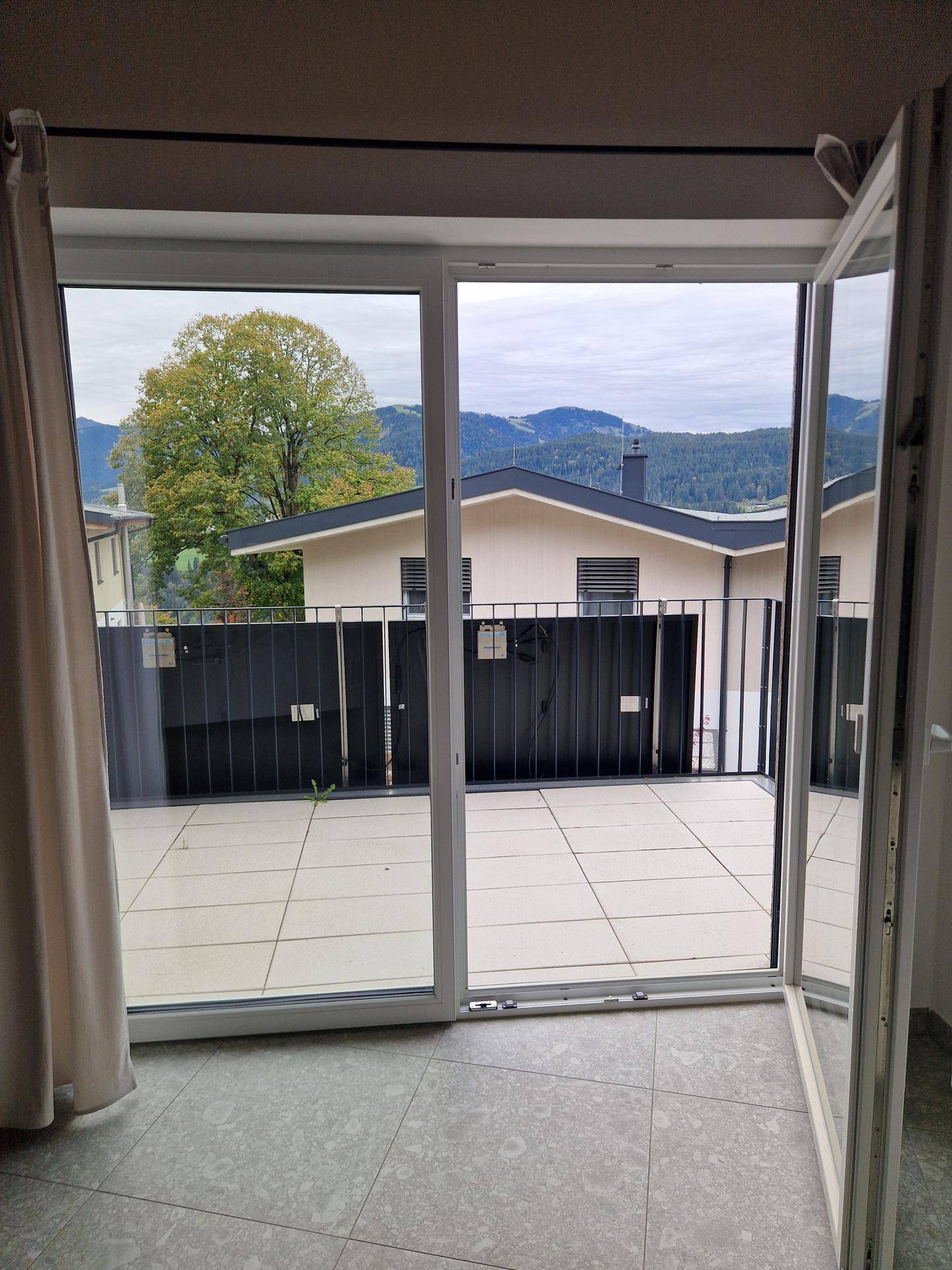Große geflieste Terrasse mit Bergblick und umliegenden Häusern, zugänglich durch eine Glastür.