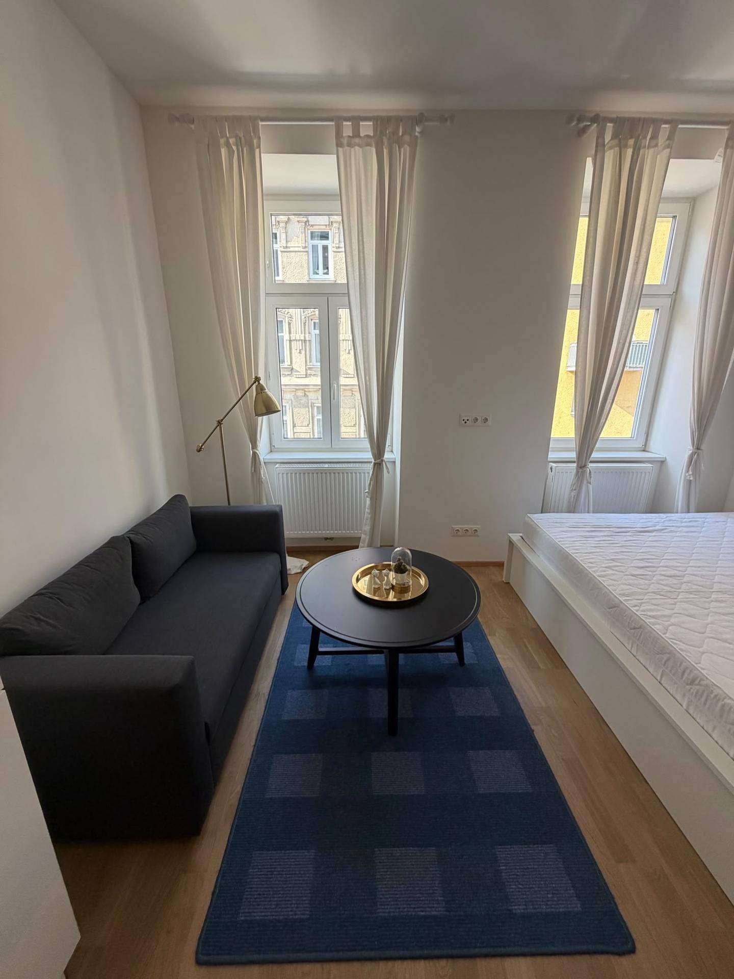 Heller Wohn- und Schlafbereich mit zwei großen Fenstern, Sofa und Bett, betont durch einen blauen Teppich.
