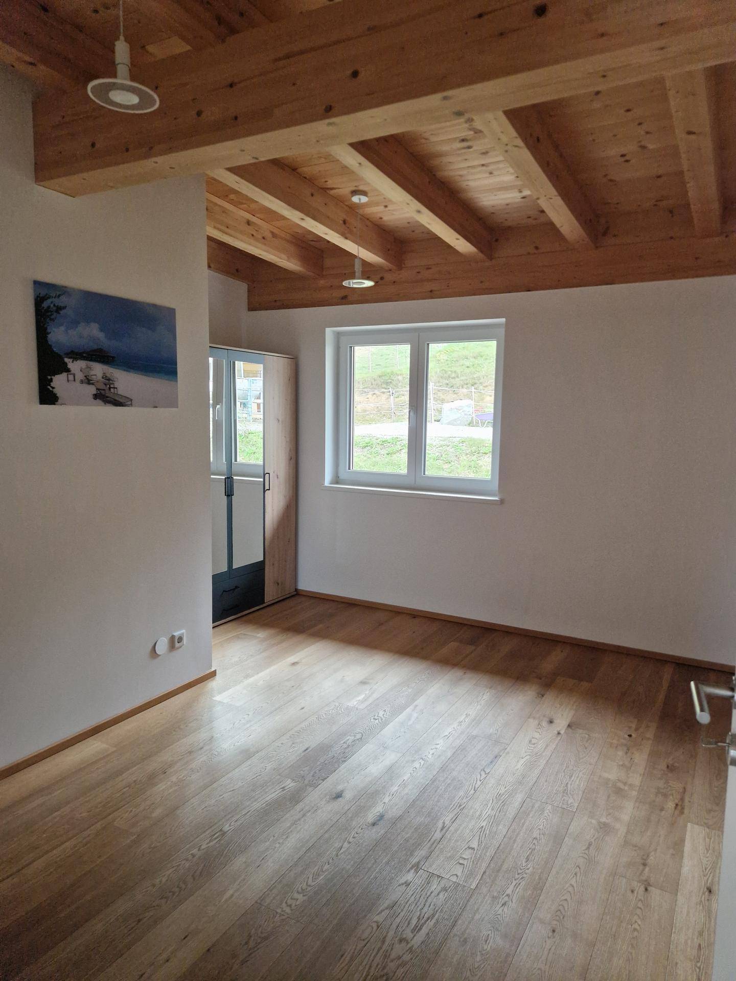 Heller Raum mit Holzbalkendecke, Dielenboden und einem Fenster, das viel Tageslicht hereinlässt.