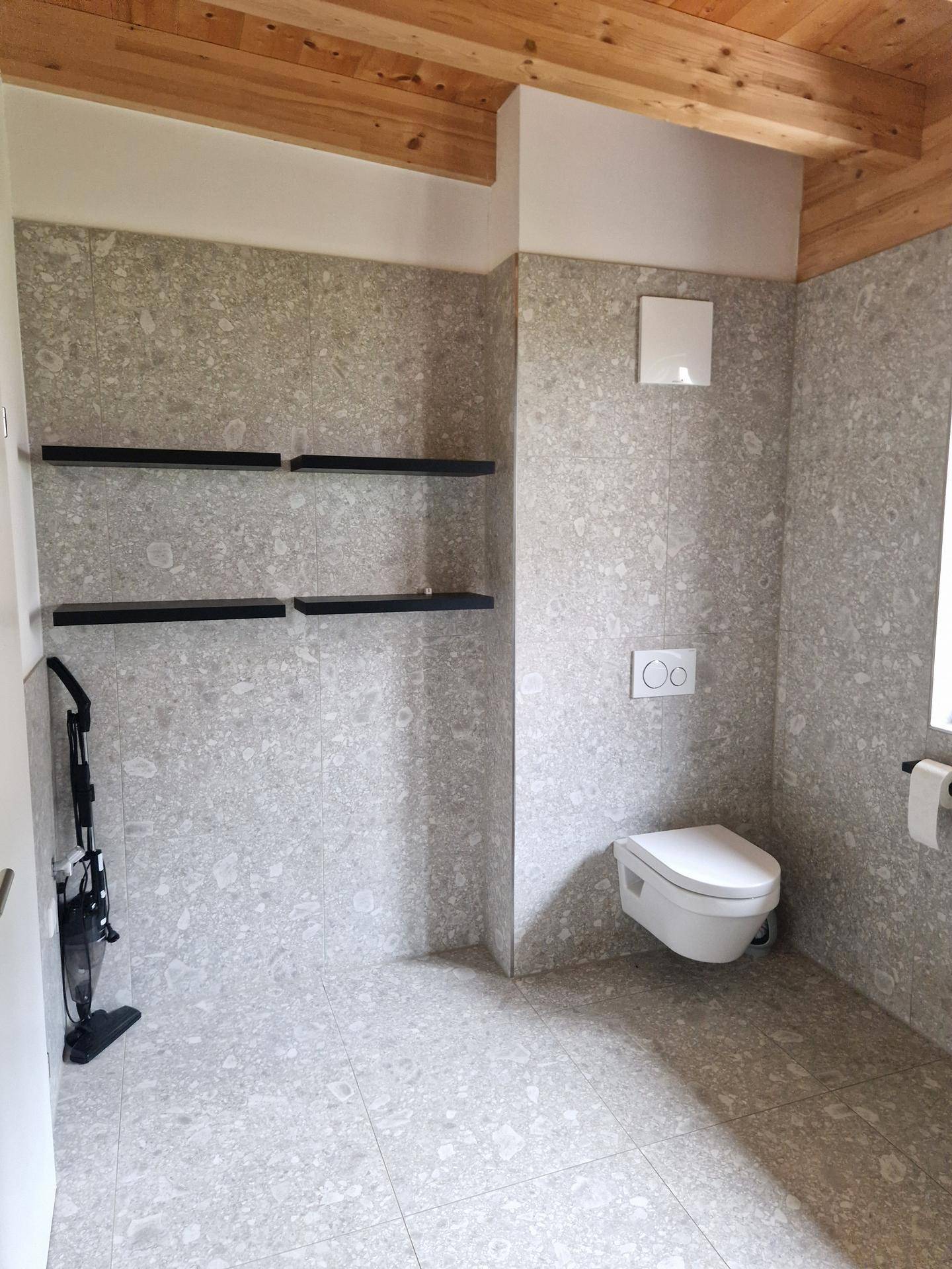 Modernes Gäste-WC mit grauen Fliesen, wandhängender Toilette und sichtbaren Holzdeckenbalken.