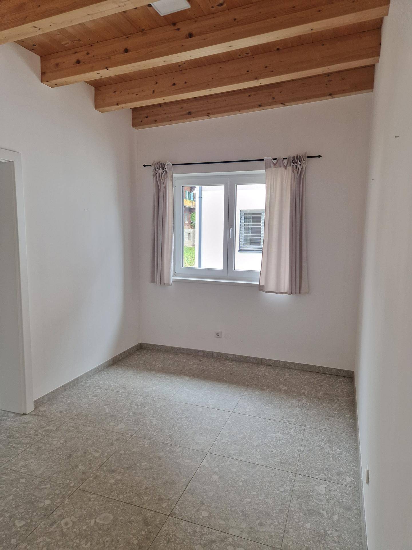 Schlafzimmer mit Fliesenboden, Holzbalkendecke und einem Fenster mit Vorhängen.