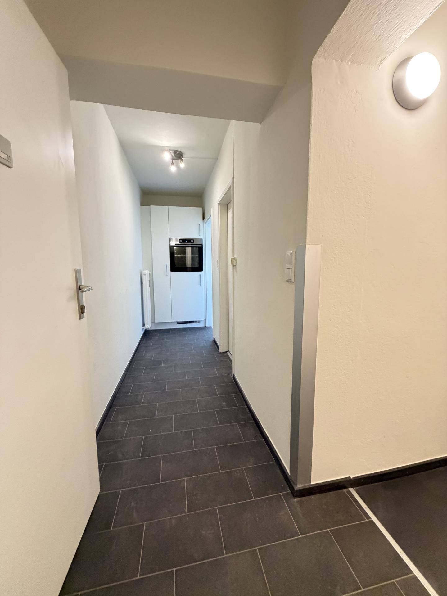 Langer Flur mit dunklem Fliesenboden, der zu einem hellen Küchenbereich führt.