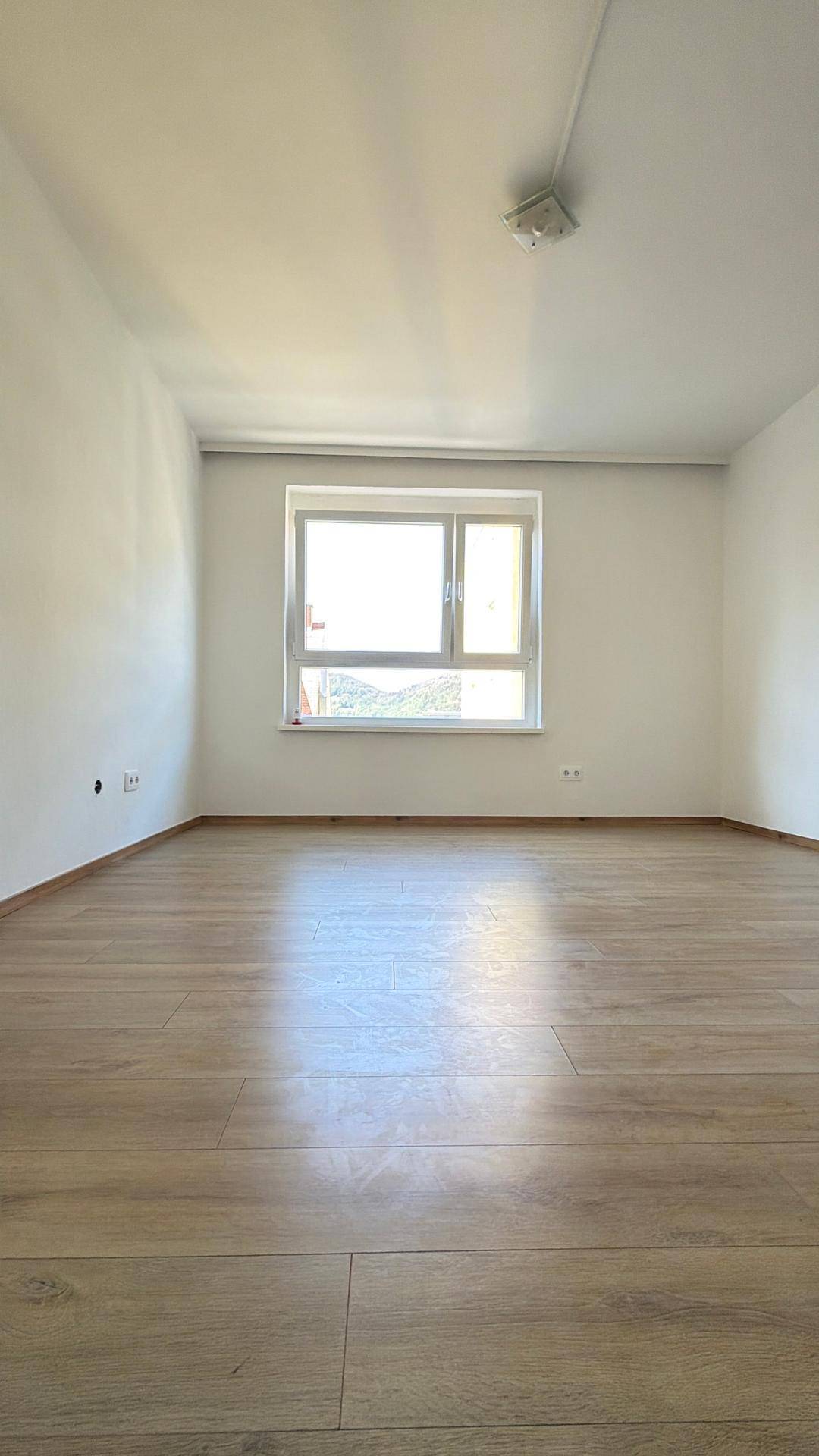 Heller, leerer Raum mit Laminatboden und einem Fenster, das viel Tageslicht hereinlässt.