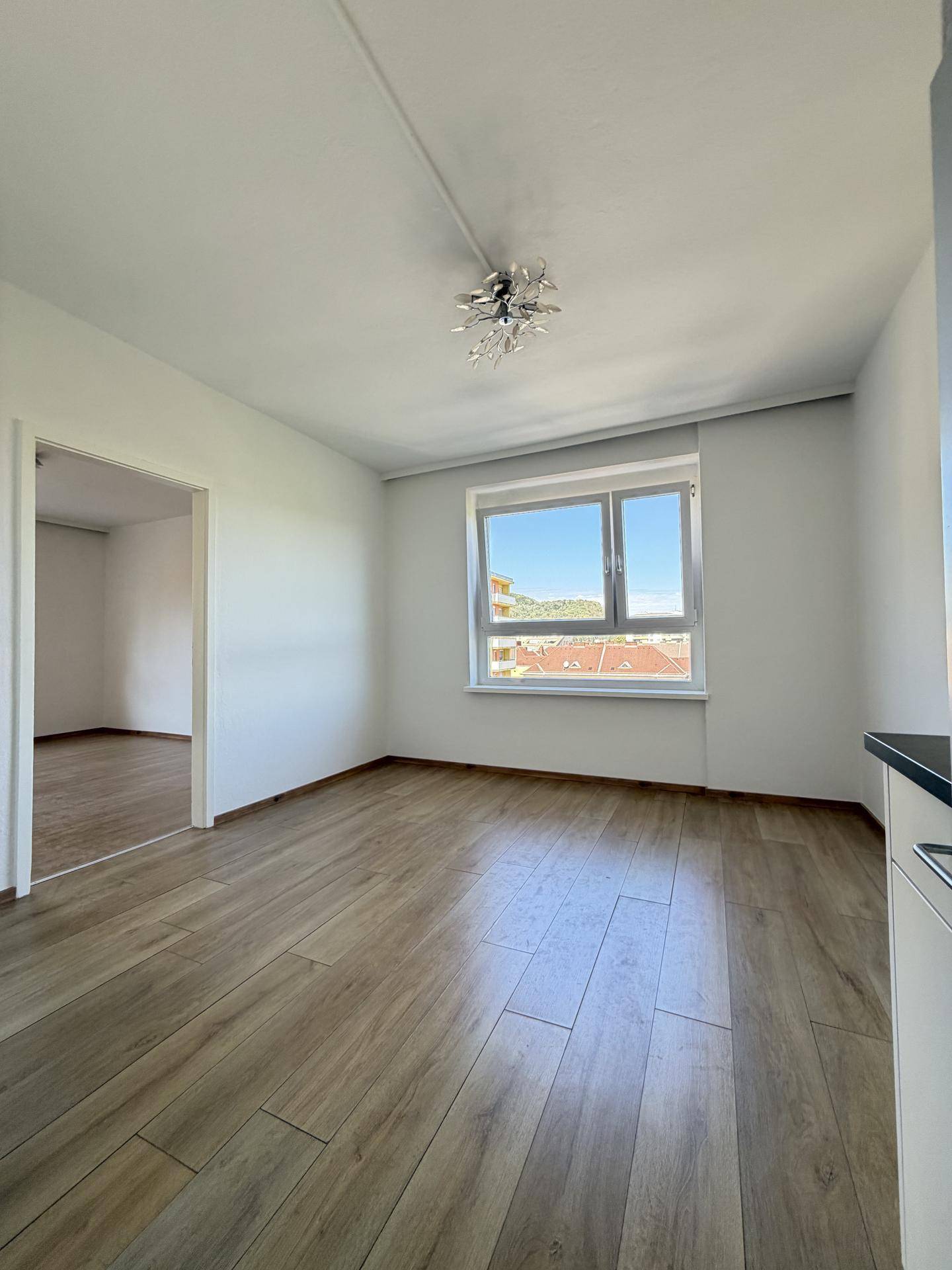 Geräumiger, heller Wohnraum mit Laminatboden, Fenster und Blick auf die Dächer der Stadt.