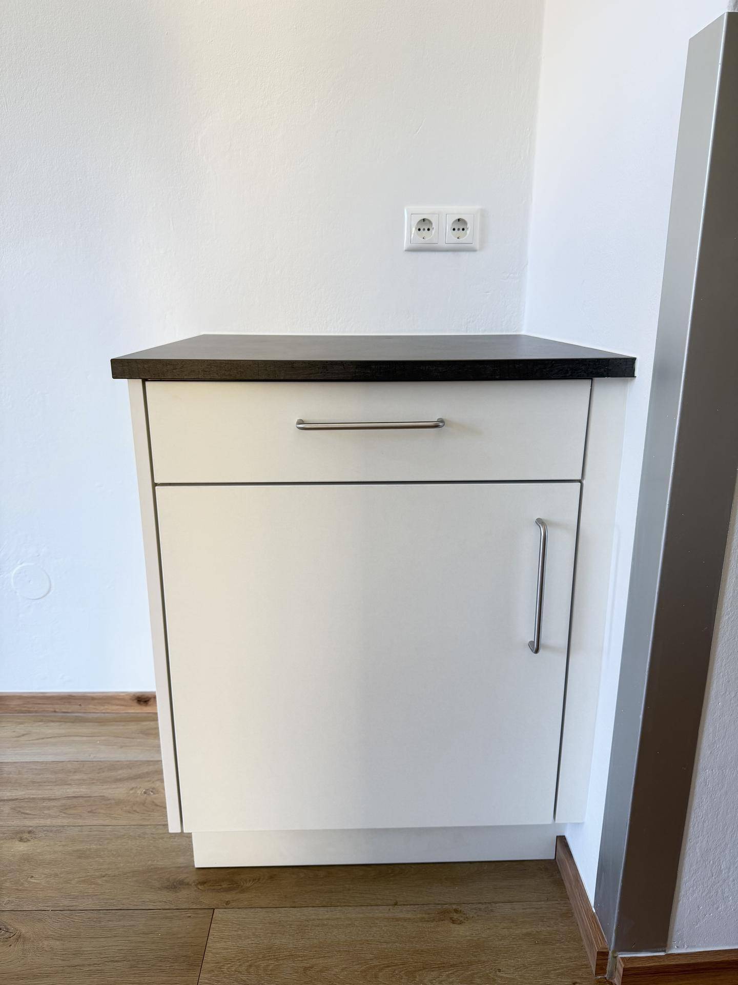 Kleiner weißer Unterschrank mit dunkler Arbeitsplatte und Schublade auf Laminatboden.