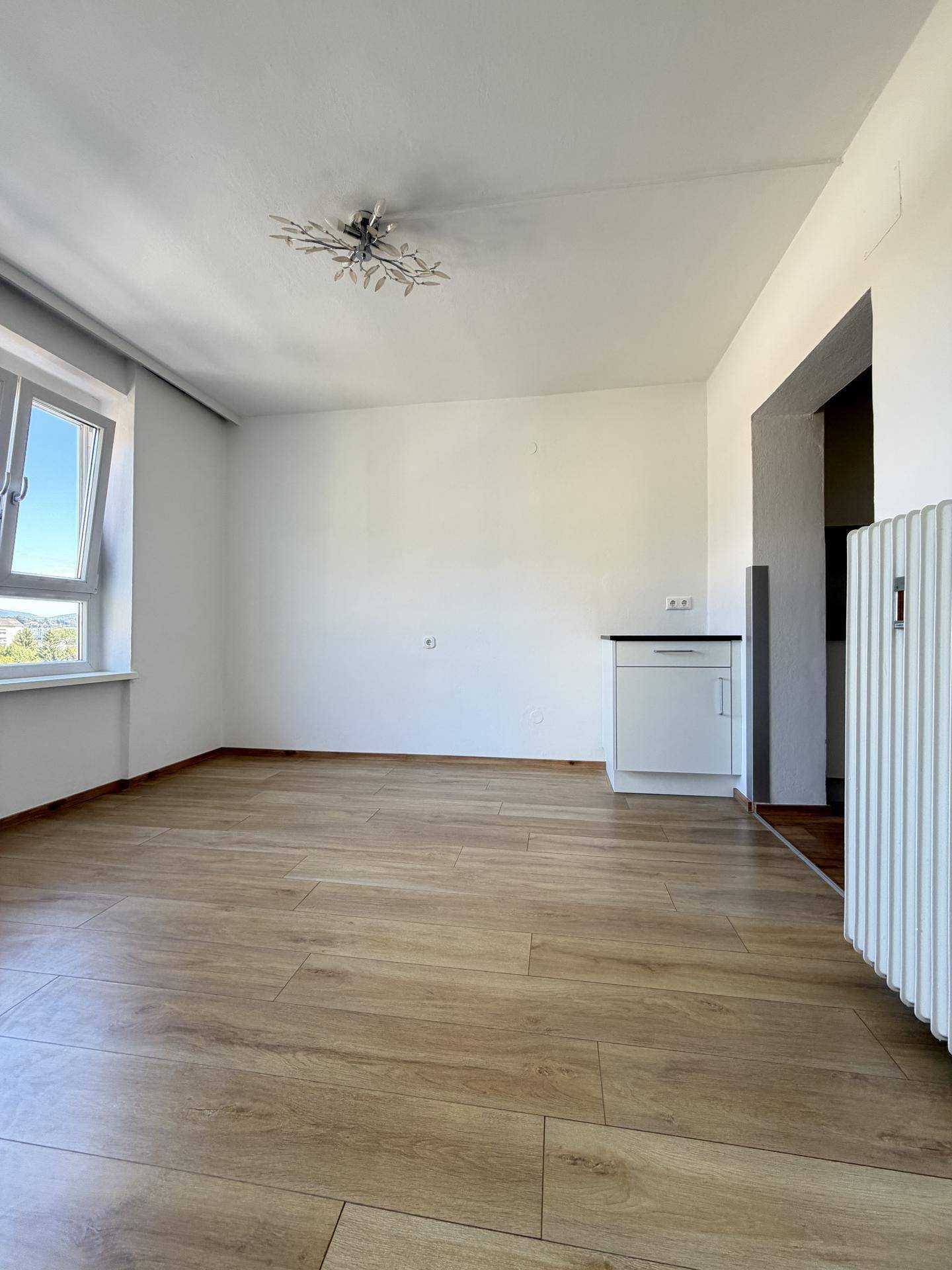 Heller Wohnraum mit Laminatboden, Heizkörper und einem kleinen Schrank unter dem Fenster.