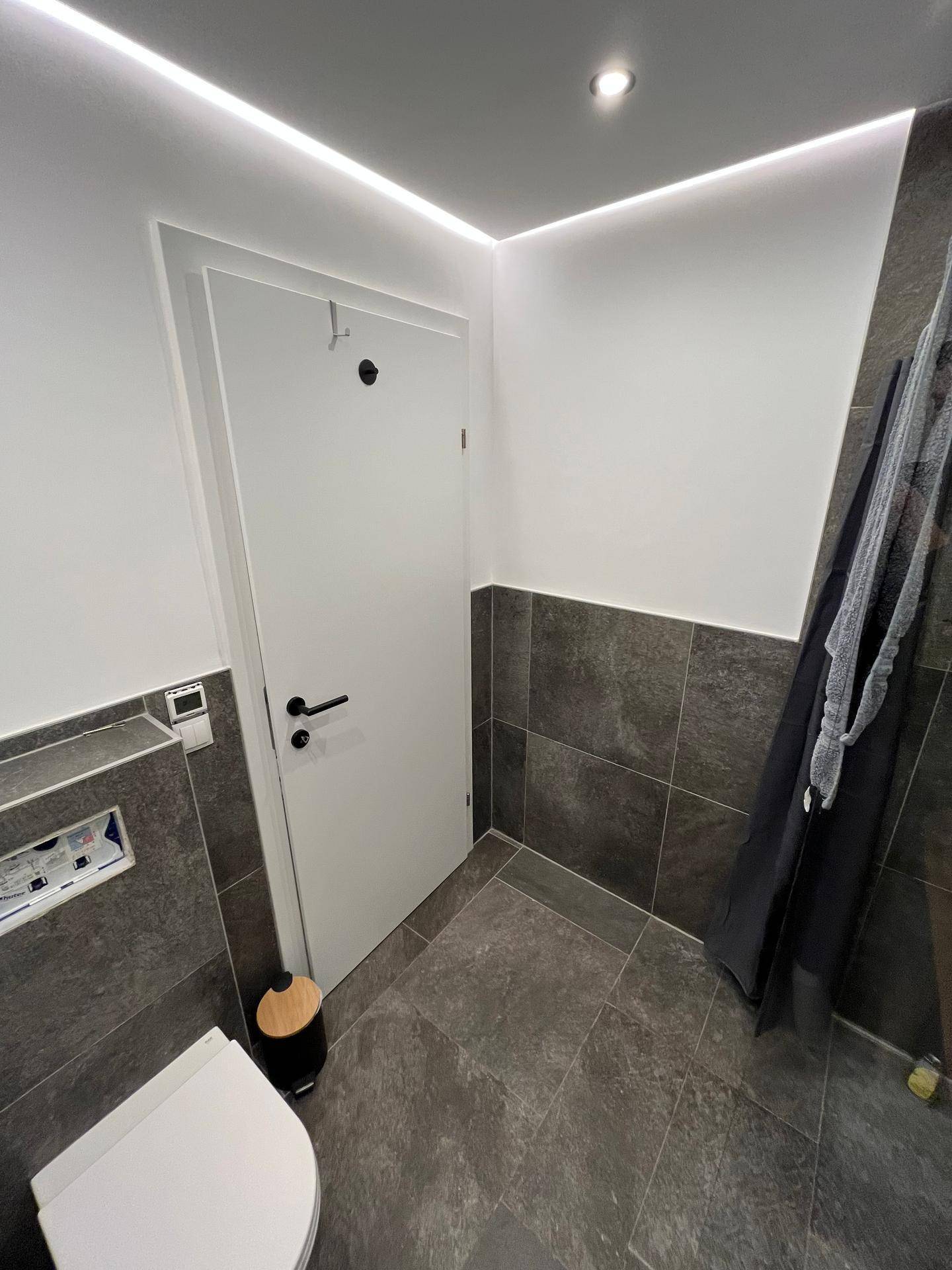 Modernes Badezimmer mit Wand-WC, dunklen Fliesen und indirekter Deckenbeleuchtung.