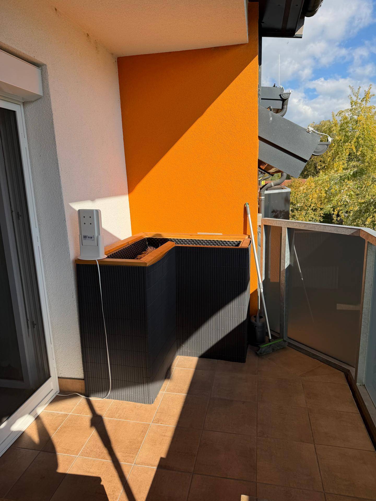 Geräumiger Balkon mit orangefarbener Akzentwand, großem Pflanzgefäß und Fliesenboden, ideal für Pflanzen.