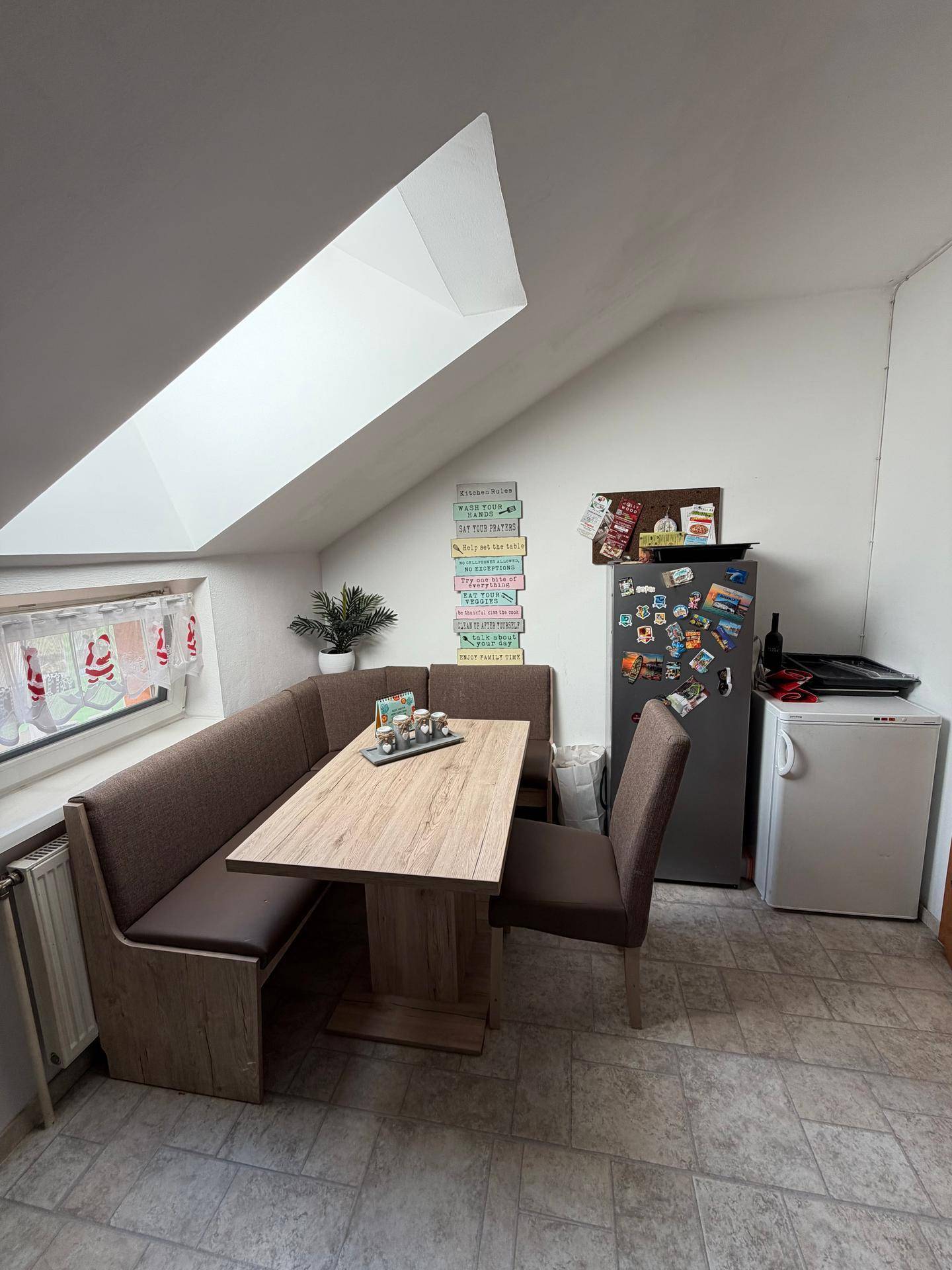 Gemütliche Wohnküche mit Eckbank, Esstisch und Kühlschrank, unter einem Dachfenster gelegen.