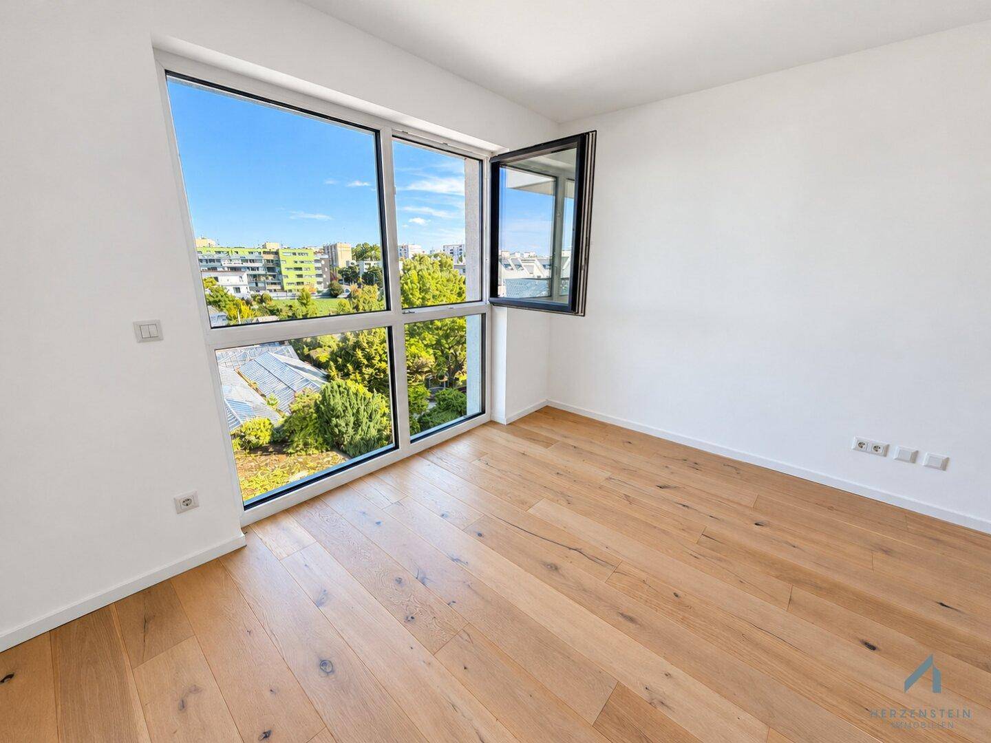 Lichtdurchflutetes Zimmer mit großen Fenstern und Blick ins Grüne, ausgestattet mit Holzparkett.