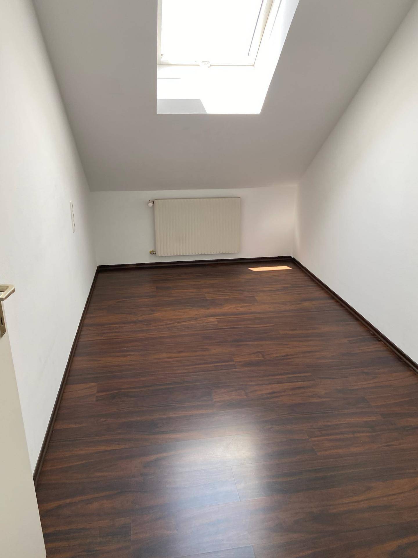 Leerer Raum im Dachgeschoss mit dunklem Laminatboden, weißen Wänden und Dachfenster.
