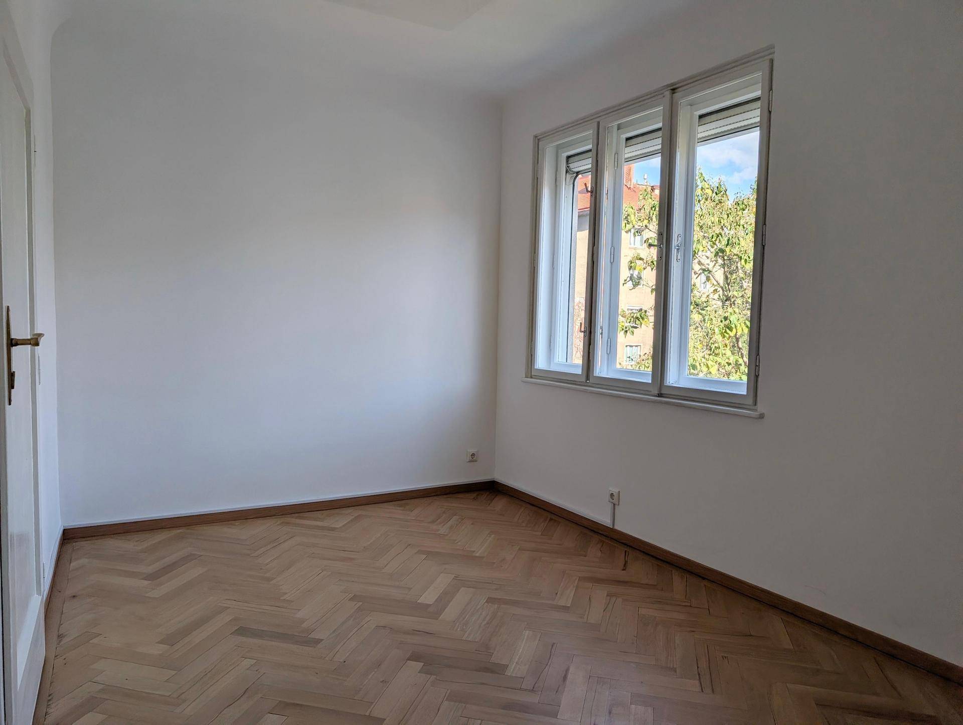 Lichtdurchflutetes Zimmer mit Fischgrätparkett und Fensterblick auf Bäume und umliegende Gebäude.