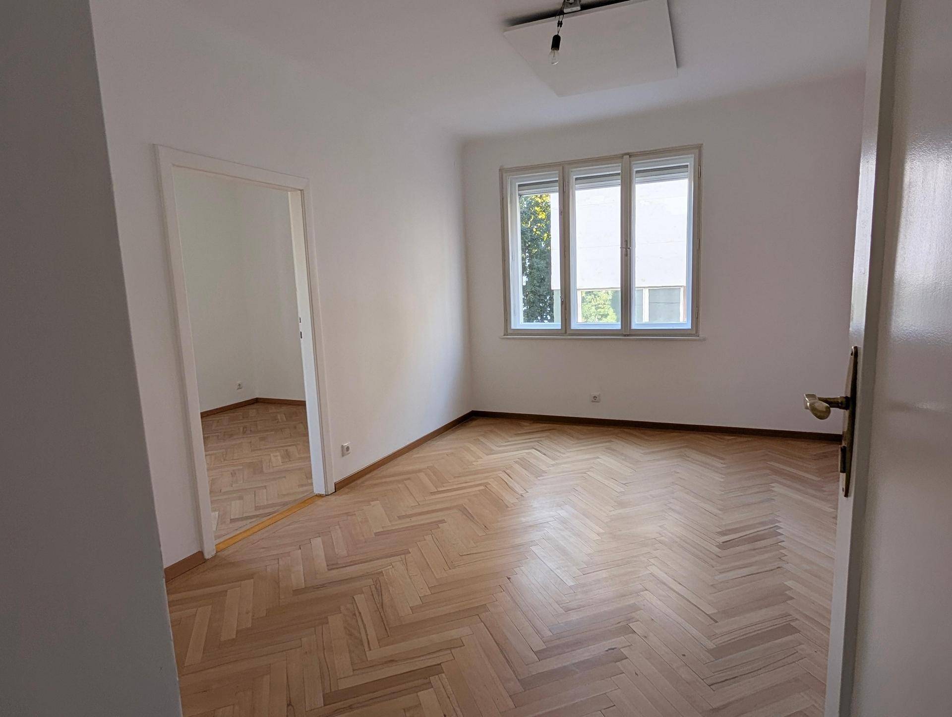 Lichtdurchflutetes Zimmer mit Fischgrätparkett und Zugang zu einem weiteren Raum, ideal für offenes Wohnen.