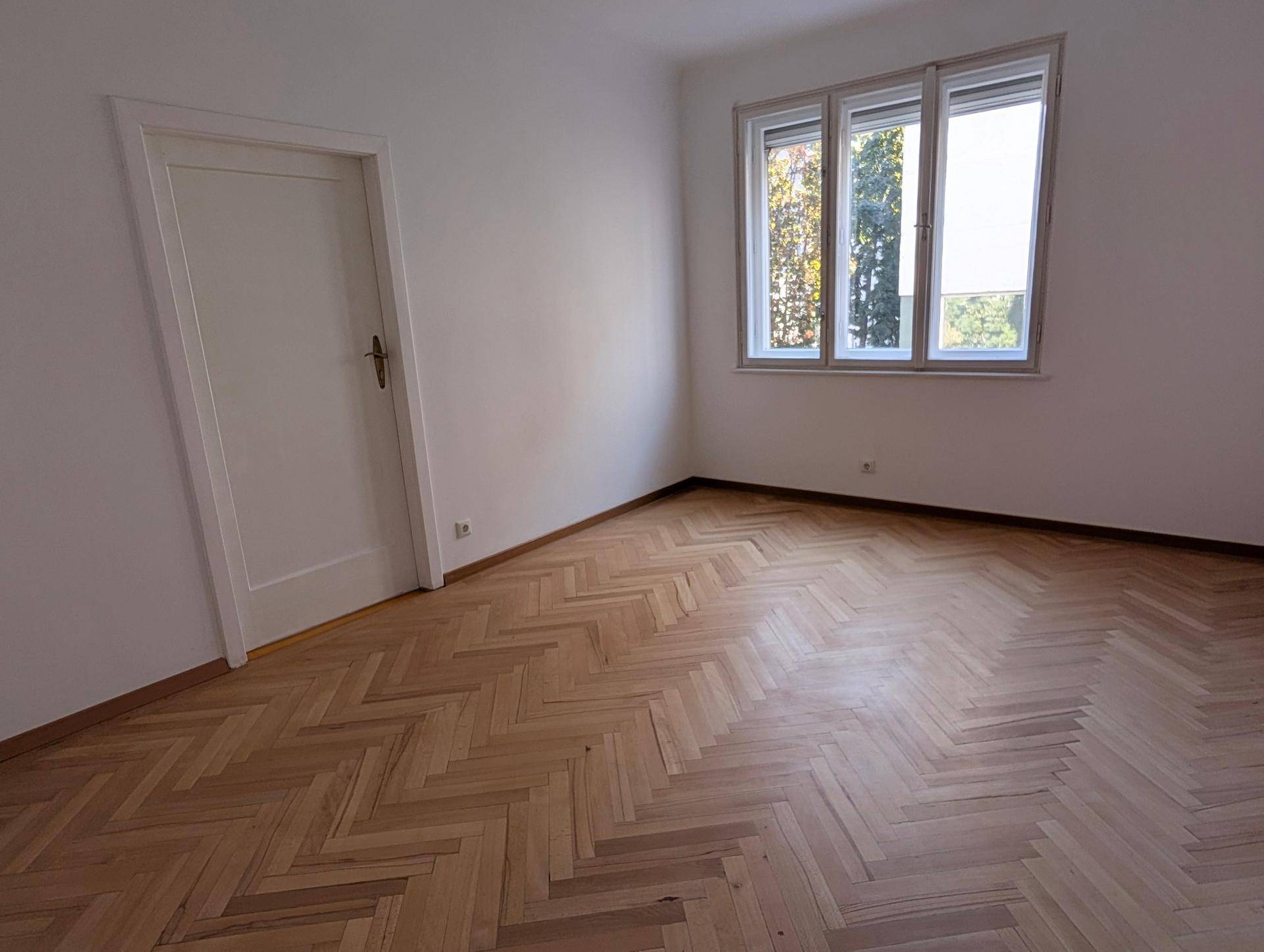 Helles Zimmer mit Fischgrätparkett, weißer Tür und Fenster mit Blick ins Grüne.