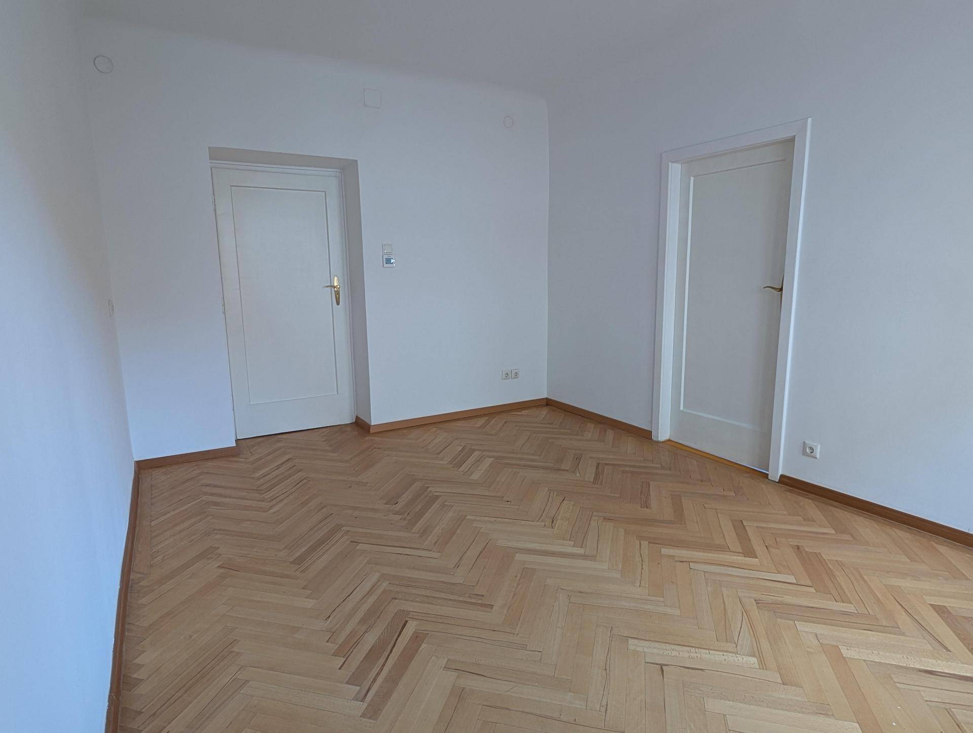 Geräumiges Zimmer mit Fischgrätparkett und zwei weißen Türen, bietet flexible Nutzungsmöglichkeiten.