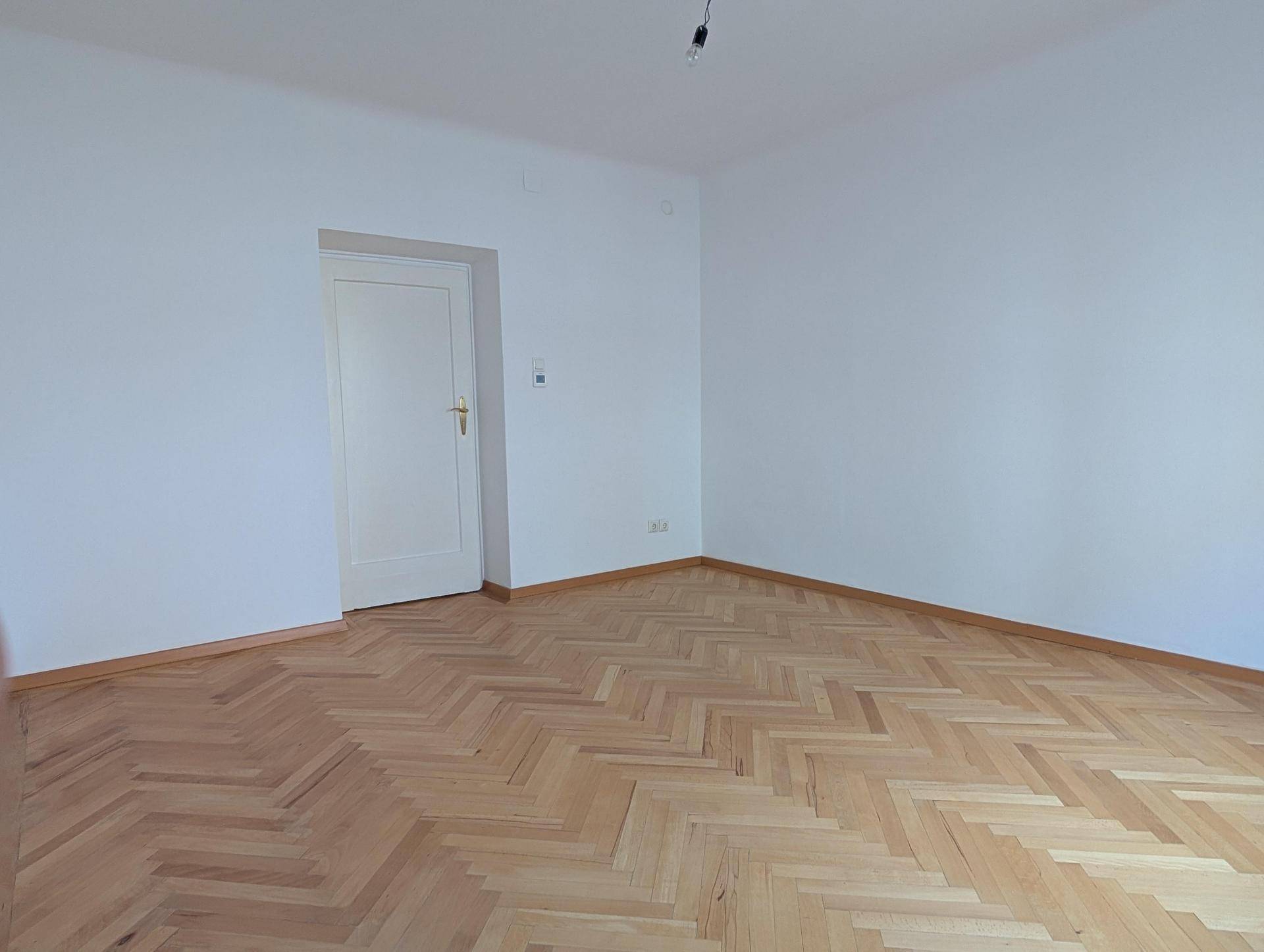 Helles Zimmer mit elegantem Fischgrätparkett und einer weißen Tür, bereit zur individuellen Gestaltung.