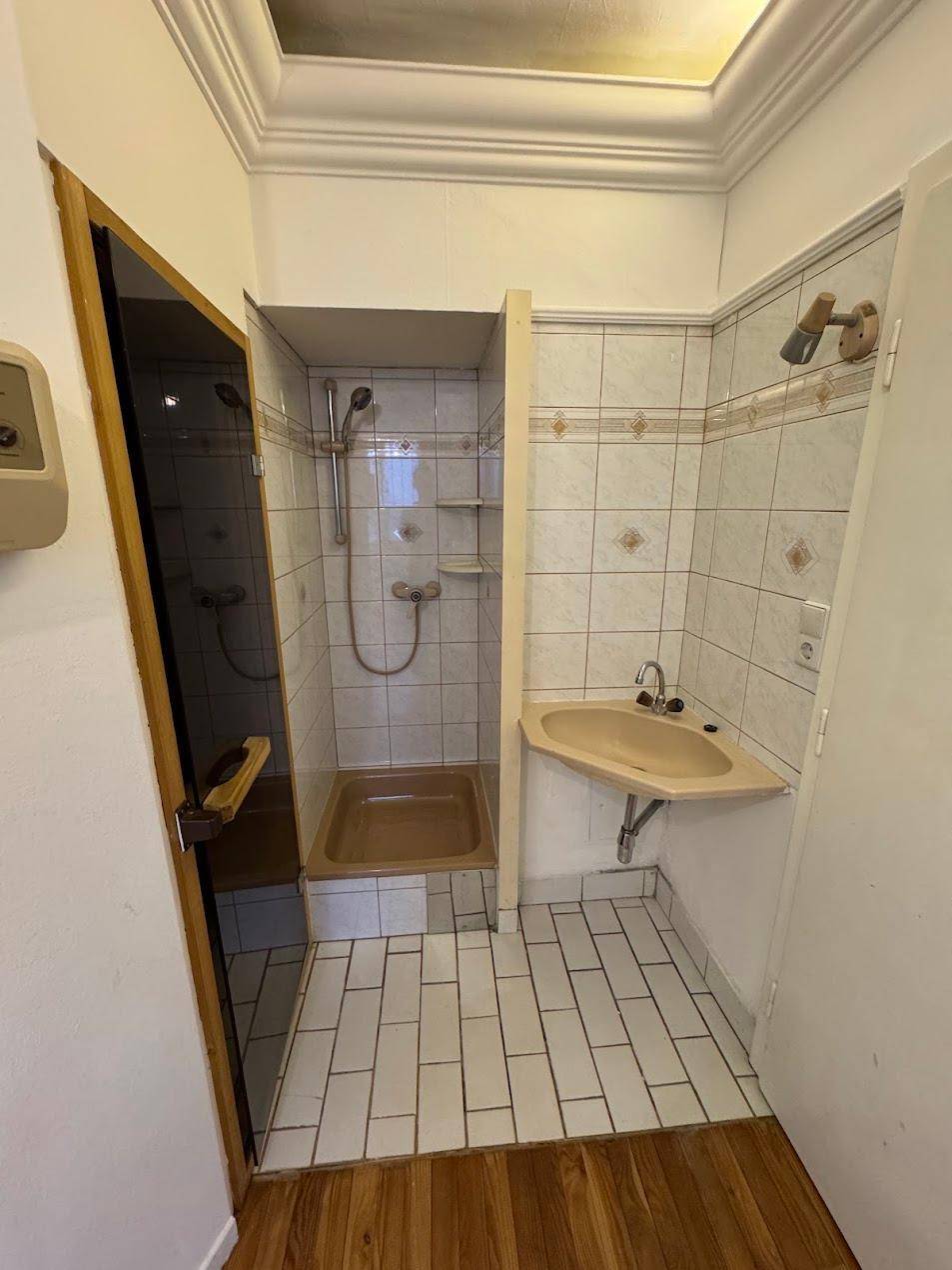 Kleines Badezimmer mit Duschkabine, Waschbecken und hellen Fliesen, in älterem Zustand.